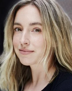 Portrait of a blonde woman with wavy hair and a neutral expression, wearing a black top.
