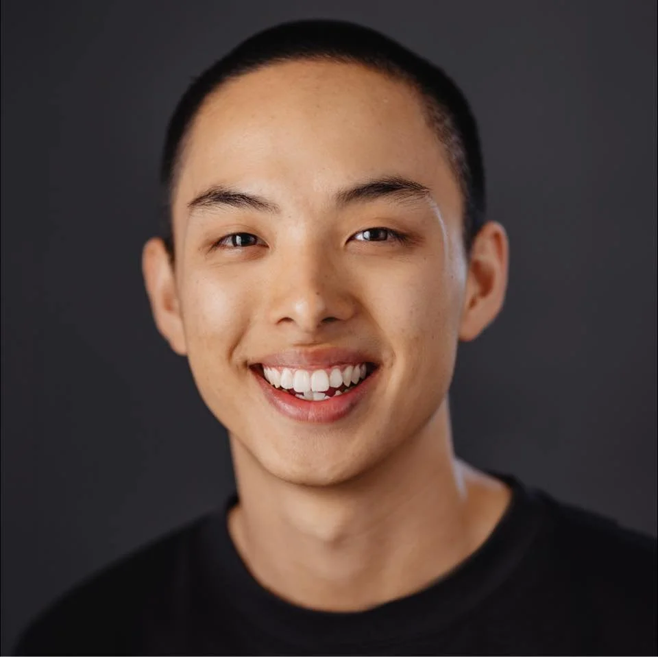 A person with short hair smiling against a dark background.