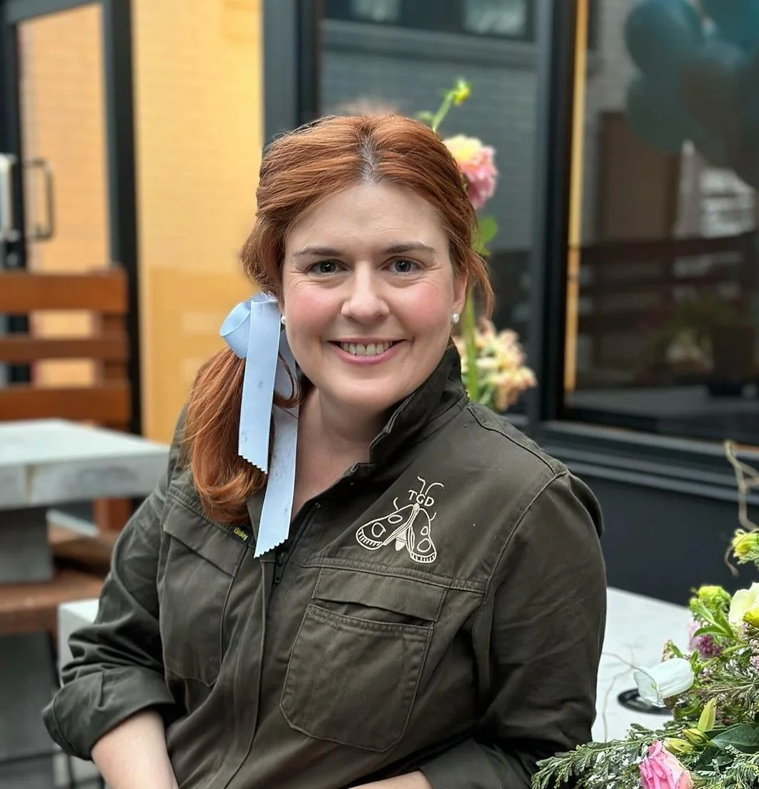 Woman with red hair smiling, wearing a green jacket with an embroidered insect design, seated in an outdoor setting with flowers and balloons in the background.
