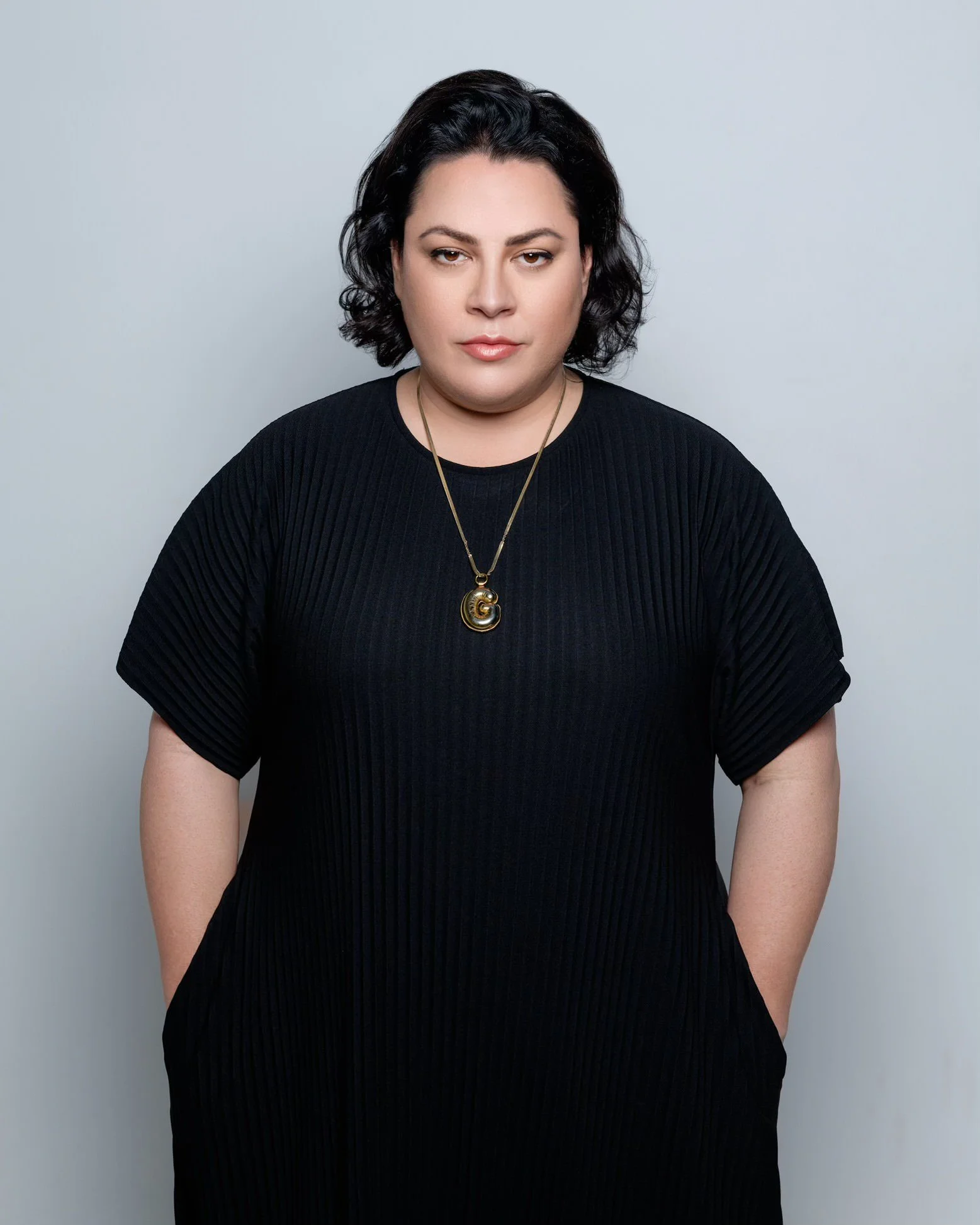 Person wearing a black ribbed dress with a pendant necklace against a gray background.