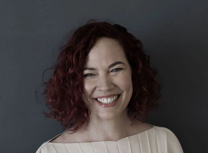 Woman with curly red hair smiling against a gray background.