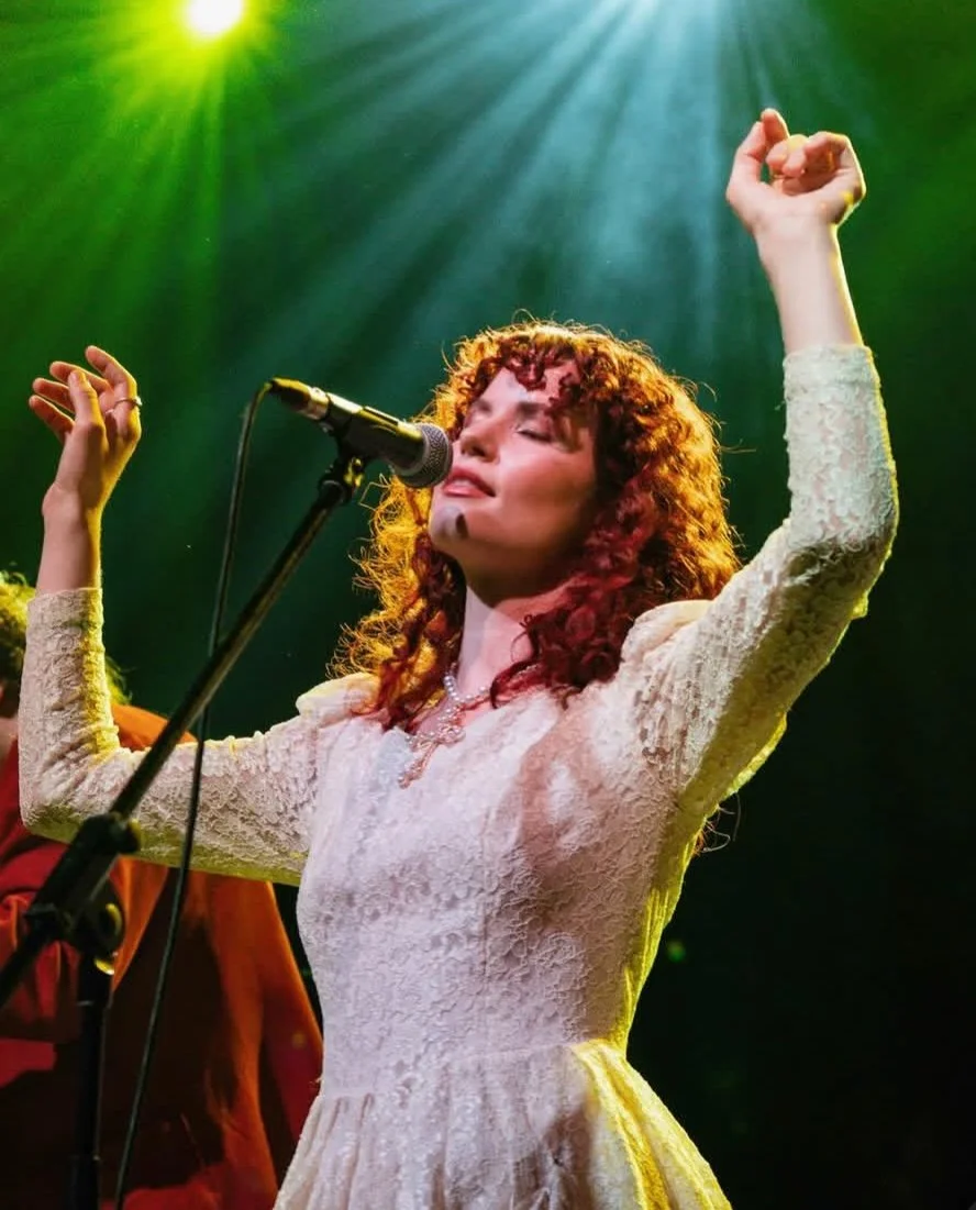 Woman with curly hair wearing a lace dress singing passionately on stage with arms raised.