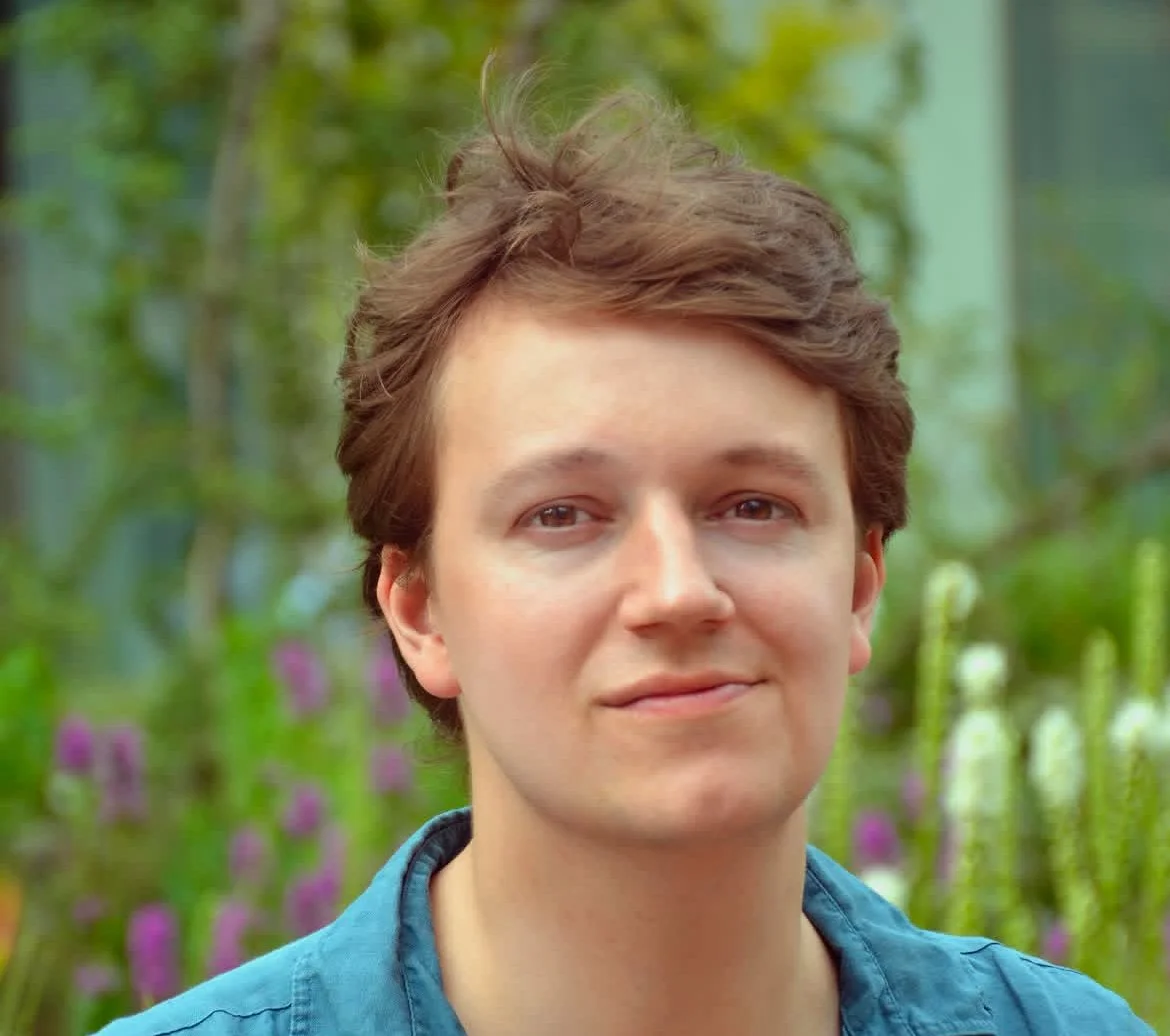 Person with light brown hair wearing a blue shirt in an outdoor setting with greenery and flowers in the background.