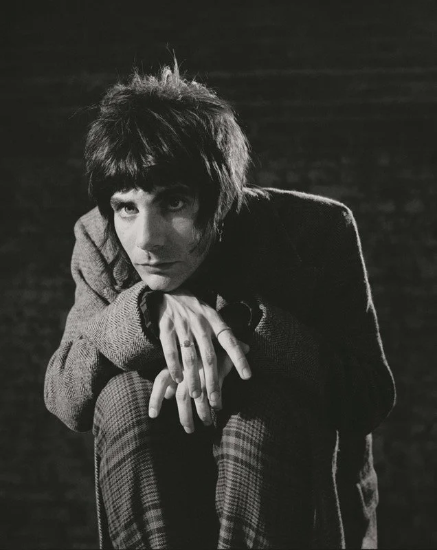 A black and white image of a person with short hair wearing a suit, leaning forward with elbows on knees and hands together, looking directly at the camera.
