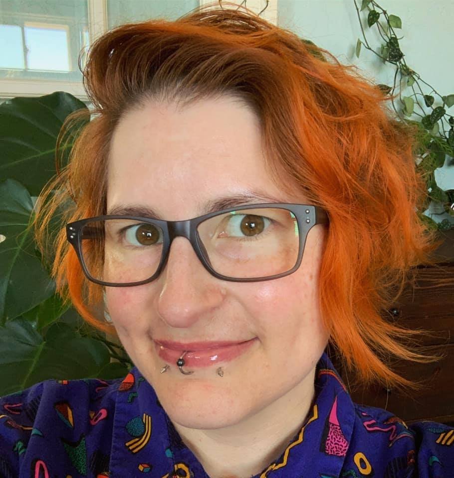 Person with glasses and orange hair, smiling, with indoor plants in the background.