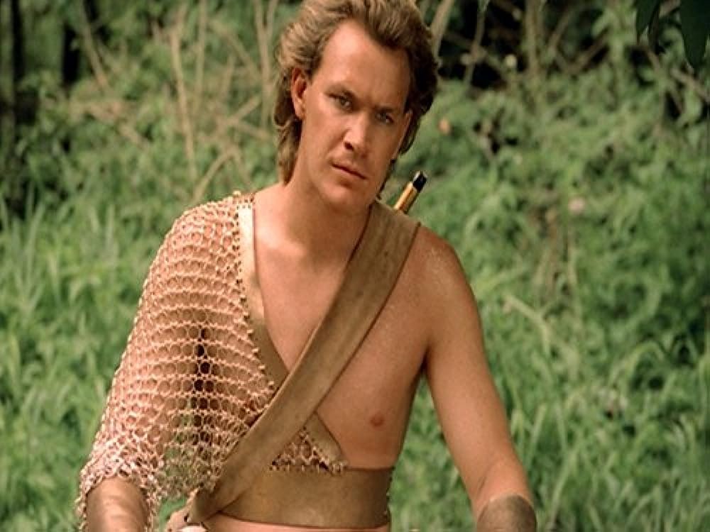 A person with messy hair wearing a mesh top sitting in front of greenery.