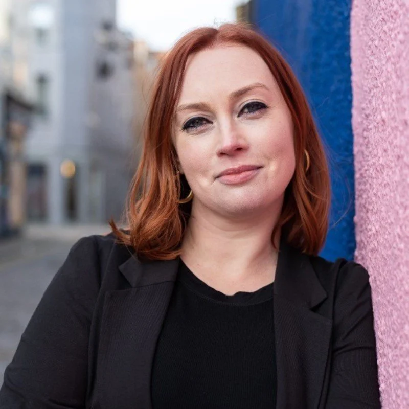 Woman with red hair leaning against a wall, wearing a black jacket and smiling slightly, with an urban background.