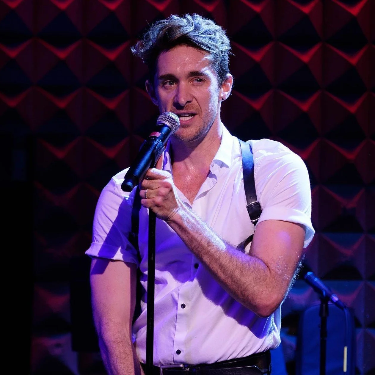 Man singing into a microphone on stage with purple-lit background.
