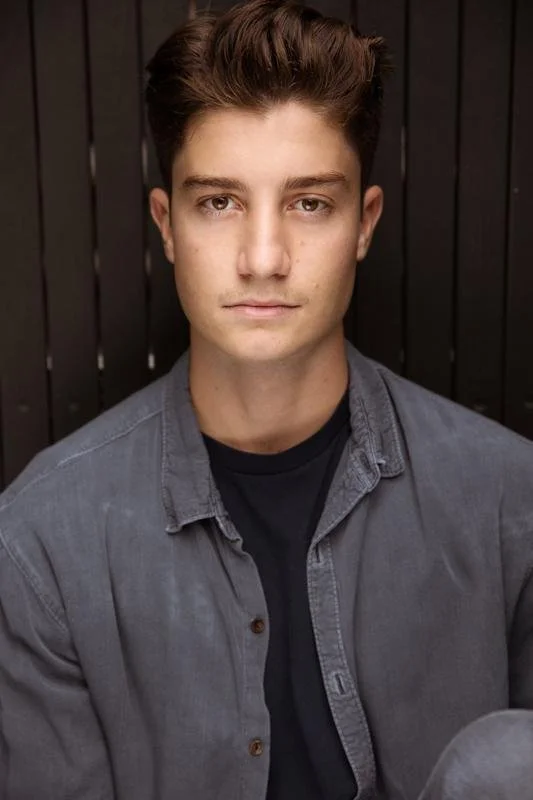 Young man with short brown hair wearing a gray shirt and black top, standing against a dark wooden background.