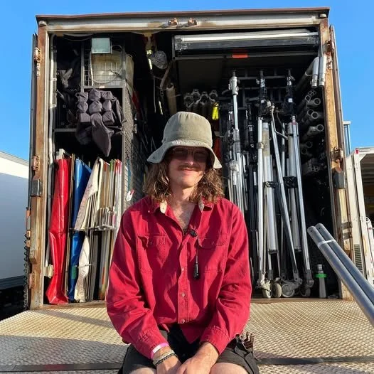 Person sitting on the back of a truck filled with equipment, wearing a red shirt and bucket hat.