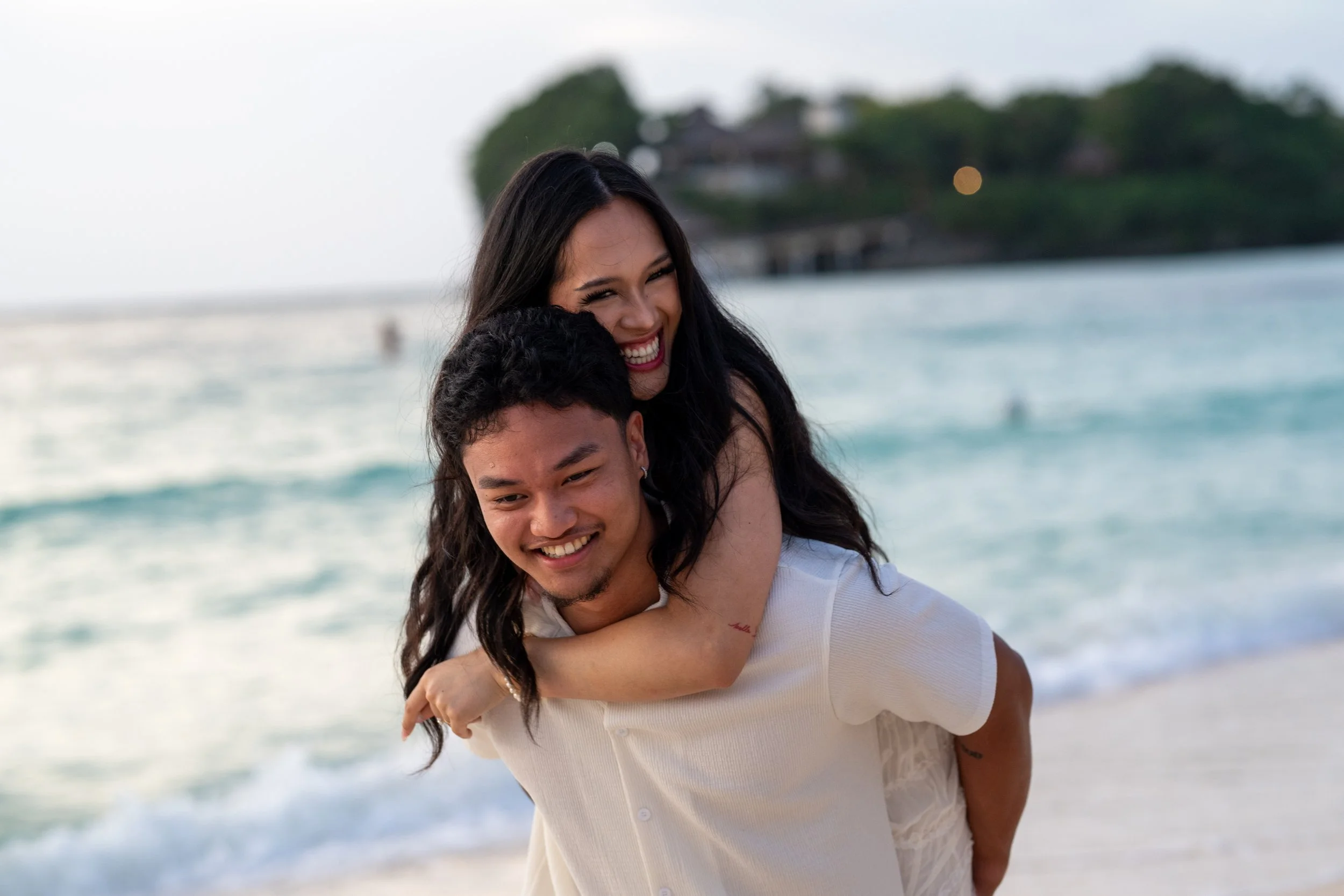 Happy couple at the beach, the woman giving the man a piggyback ride, ocean waves, and a pier in the background.