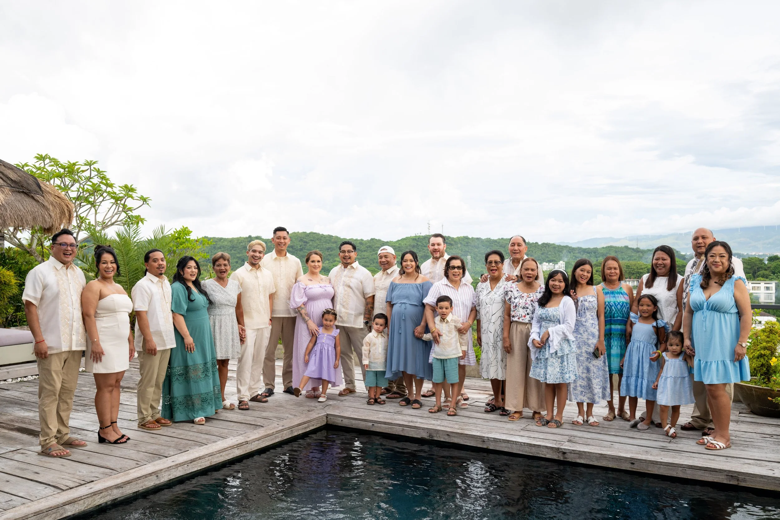 Group of family and friends dressed in light-colored and pastel clothing, standing on a wooden deck near a pool with a scenic green mountain view in the background.