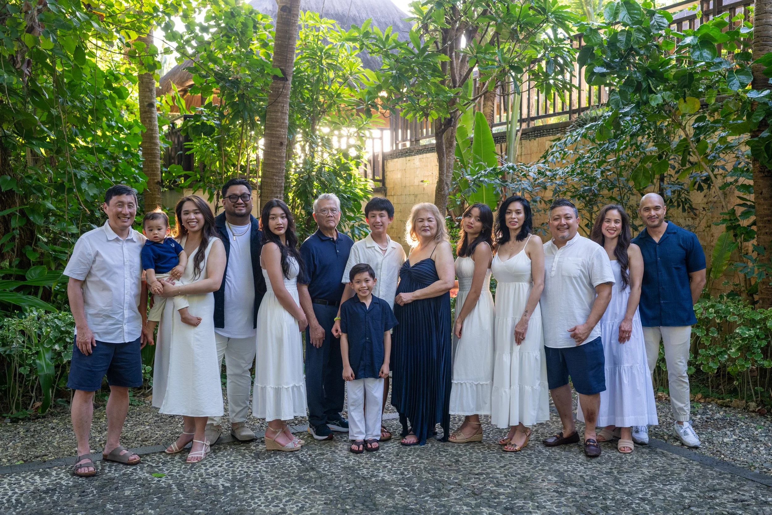 A diverse group of seventeen people, including children, adults, and seniors, smiling and posing outdoors in a lush garden with large green tropical plants and trees.
