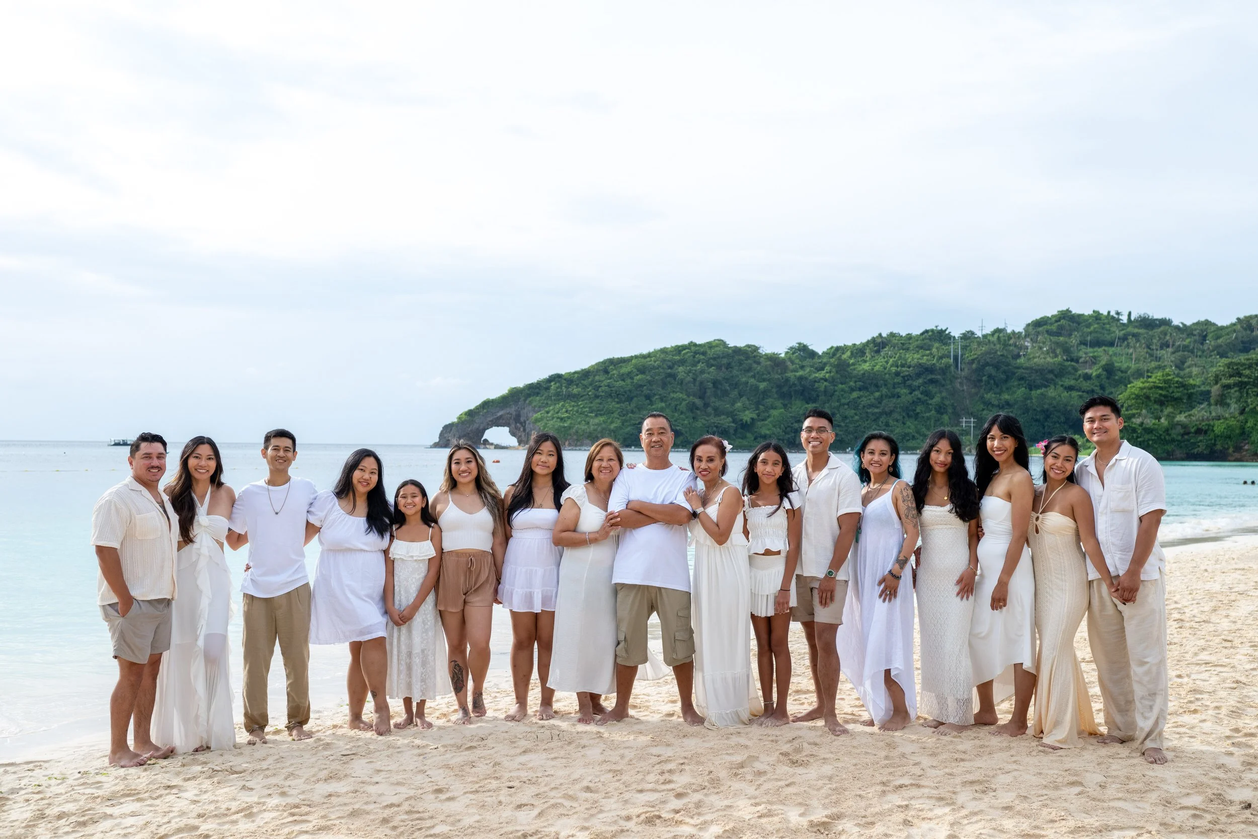 A large group of people standing on a sandy beach with a green hill and ocean in the background, all dressed in white and beige clothing.