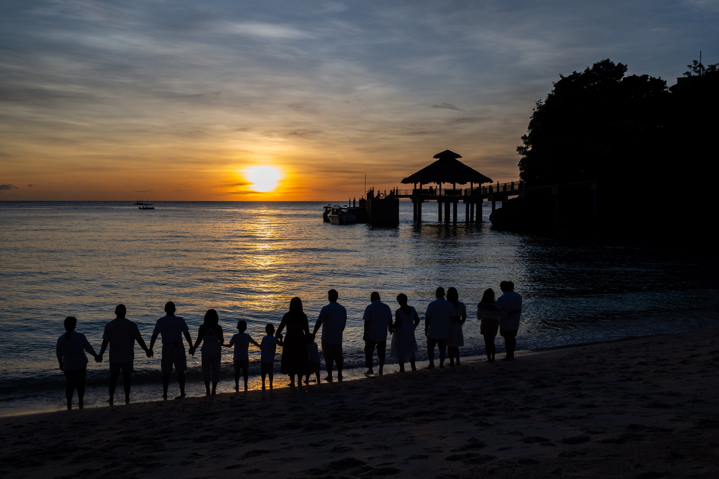 Silhouetted family holding hands along a beach at sunset with a pier and a thatched-roof structure over the water.