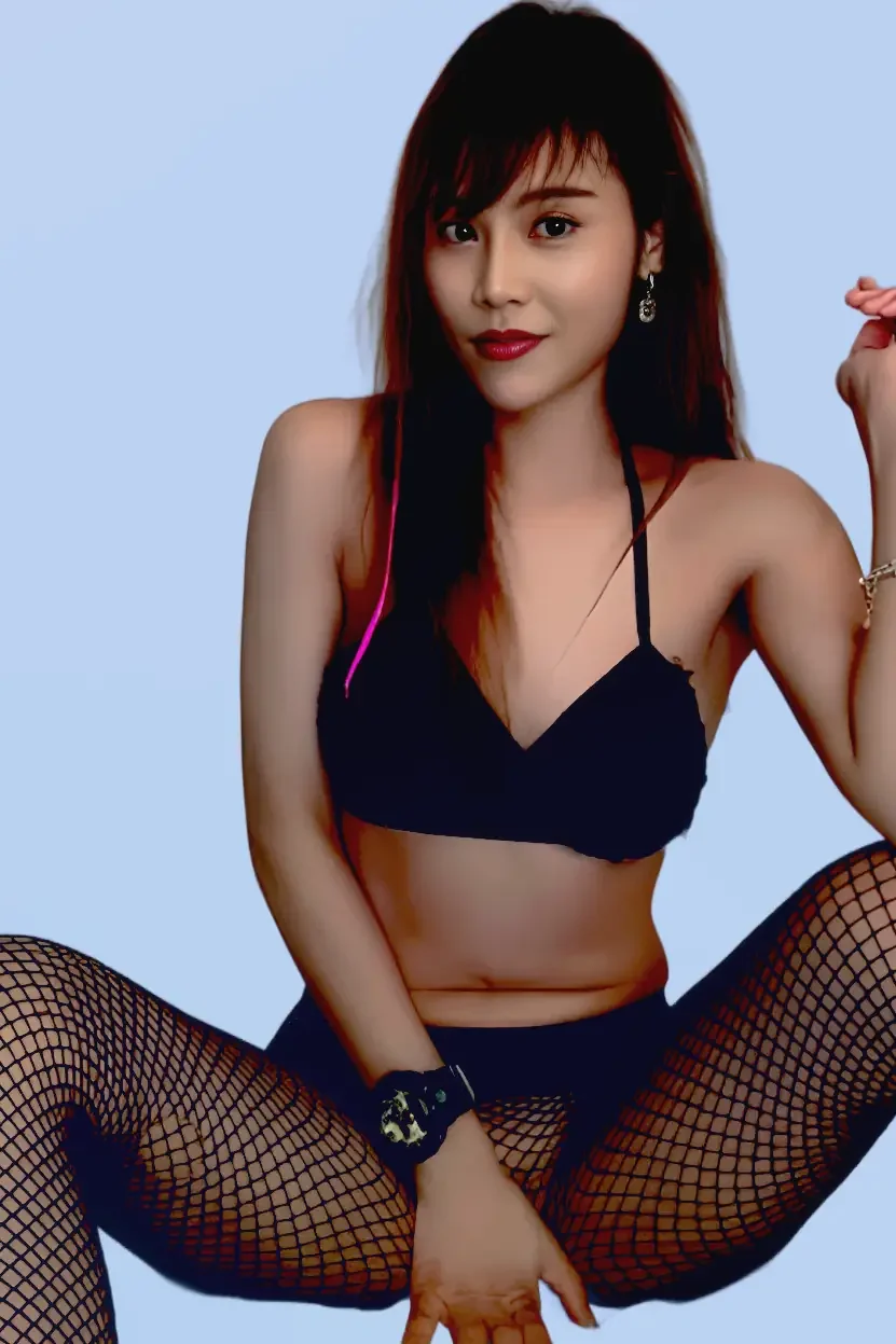 A young woman with red hair and bangs, wearing a black crop top, fishnet stockings, and jewelry, sitting on a bed with a light green background.