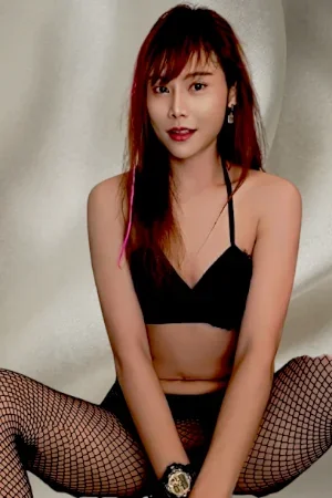 A young woman with red hair and bangs, wearing a black crop top, fishnet stockings, and jewelry, sitting on a white silk sheet.