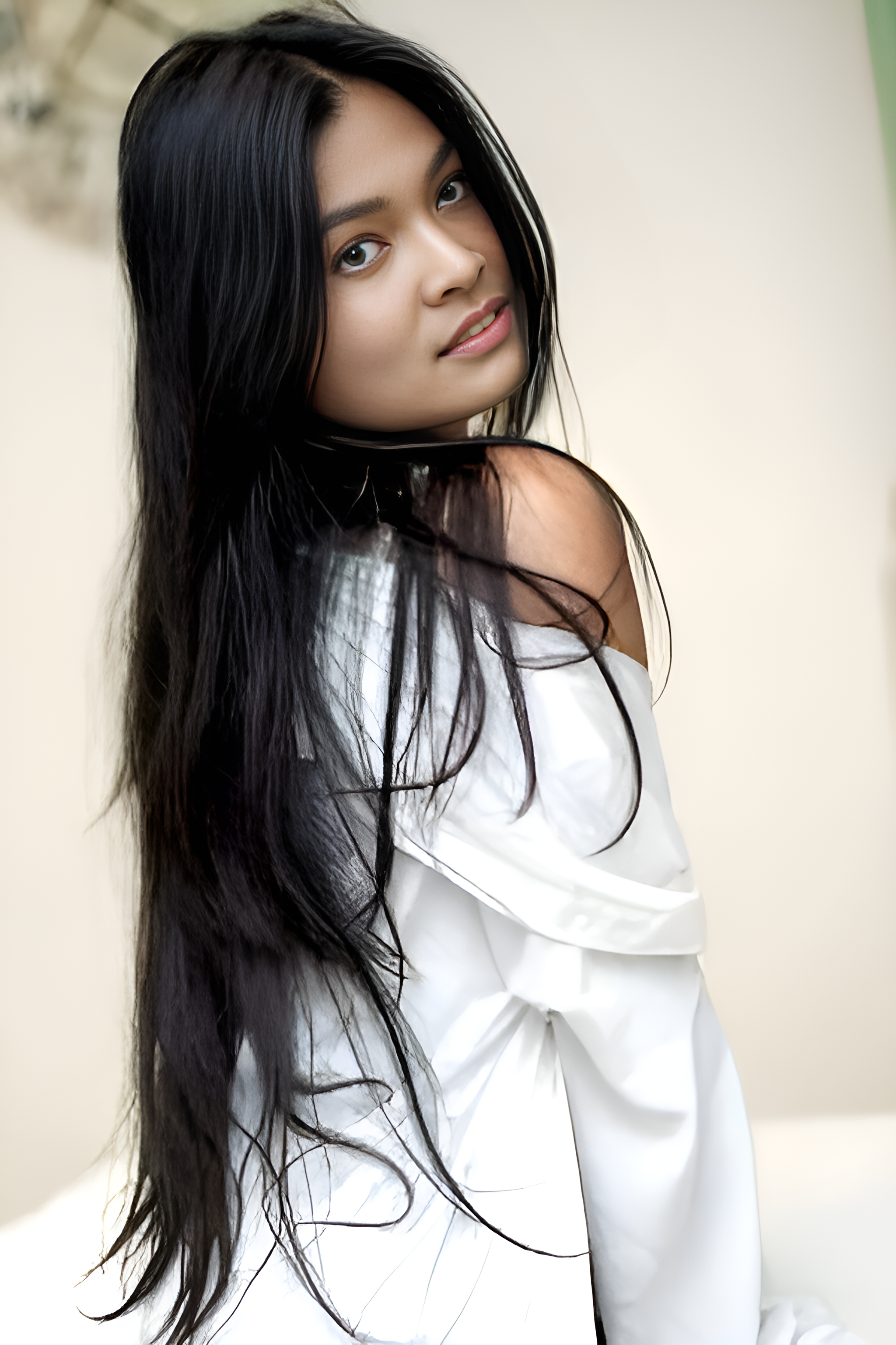 A young woman with long black hair and light skin, wearing a white top, looks over her shoulder at the camera.