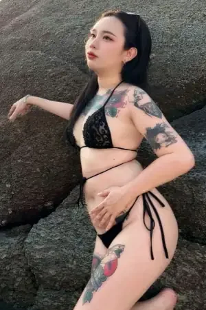 Эскорт-девушка из Паттайи по имени Фай, украшенная татуировками, отдыхает на камне, сочетая свой уникальный стиль с безмятежной атмосферой на открытом воздухе.