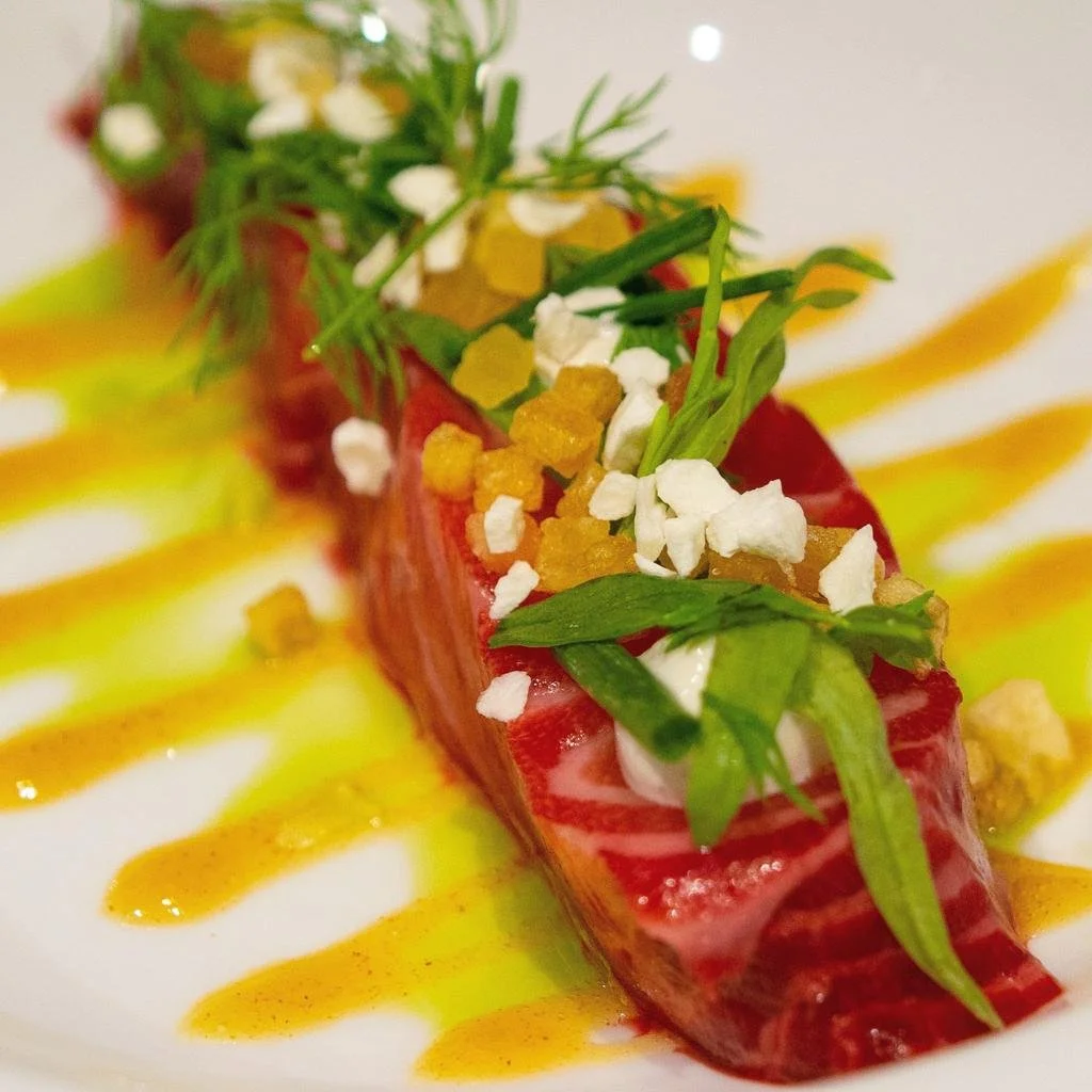 Een gerecht met rauwe tonijn, gegarneerd met groene kruiden, krokante witte stukjes, groene bladgroenten, en een saus met gele en oranje tinten.