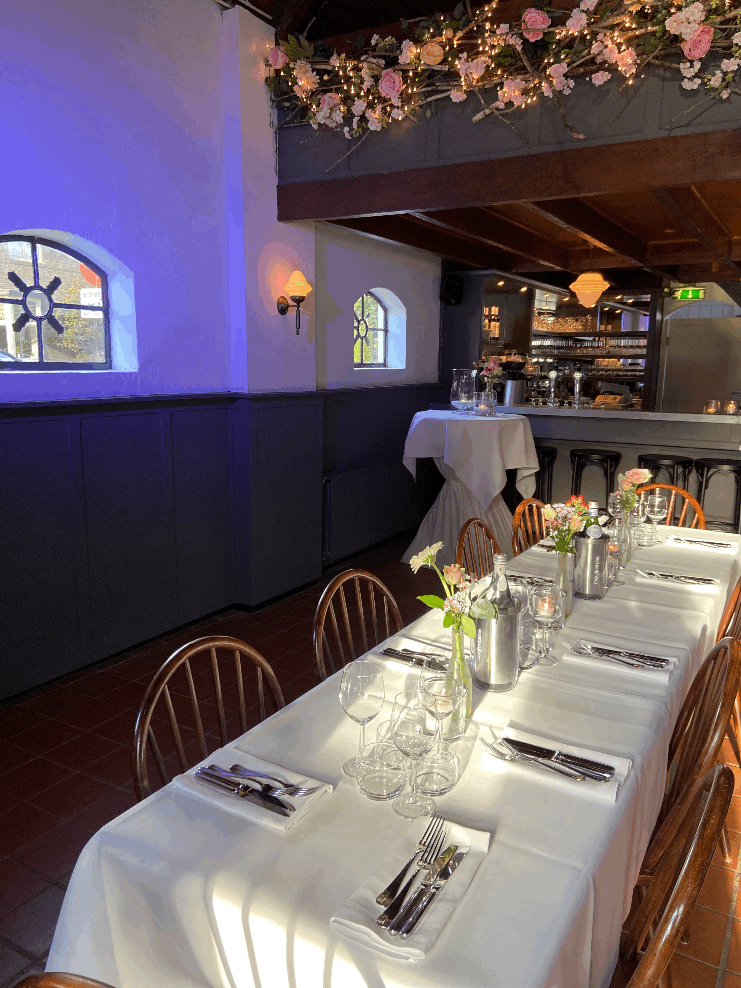 Een mooi gedekte tafel in een restaurant met witte tafelkleed, glazen, bestek, wijnflessen en bloemen, onder een plafond met bloemen en lichtjes.