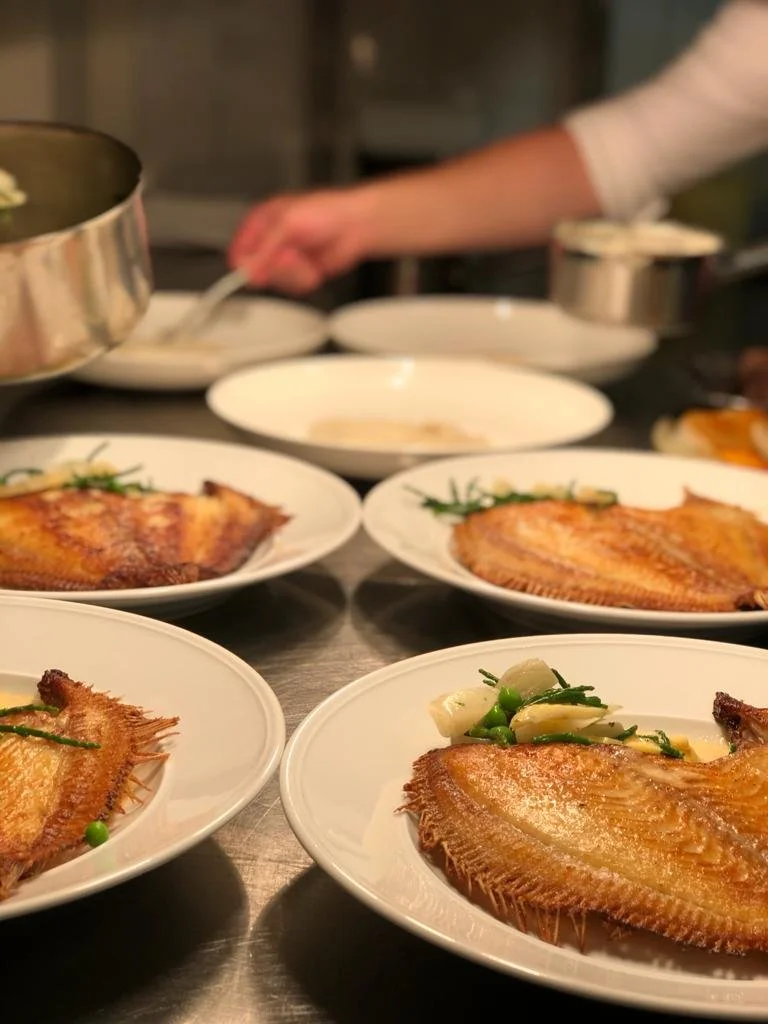 Gerechten met gebakken vis op witte borden, terwijl een persoon in de achtergrond gerechten serveert.