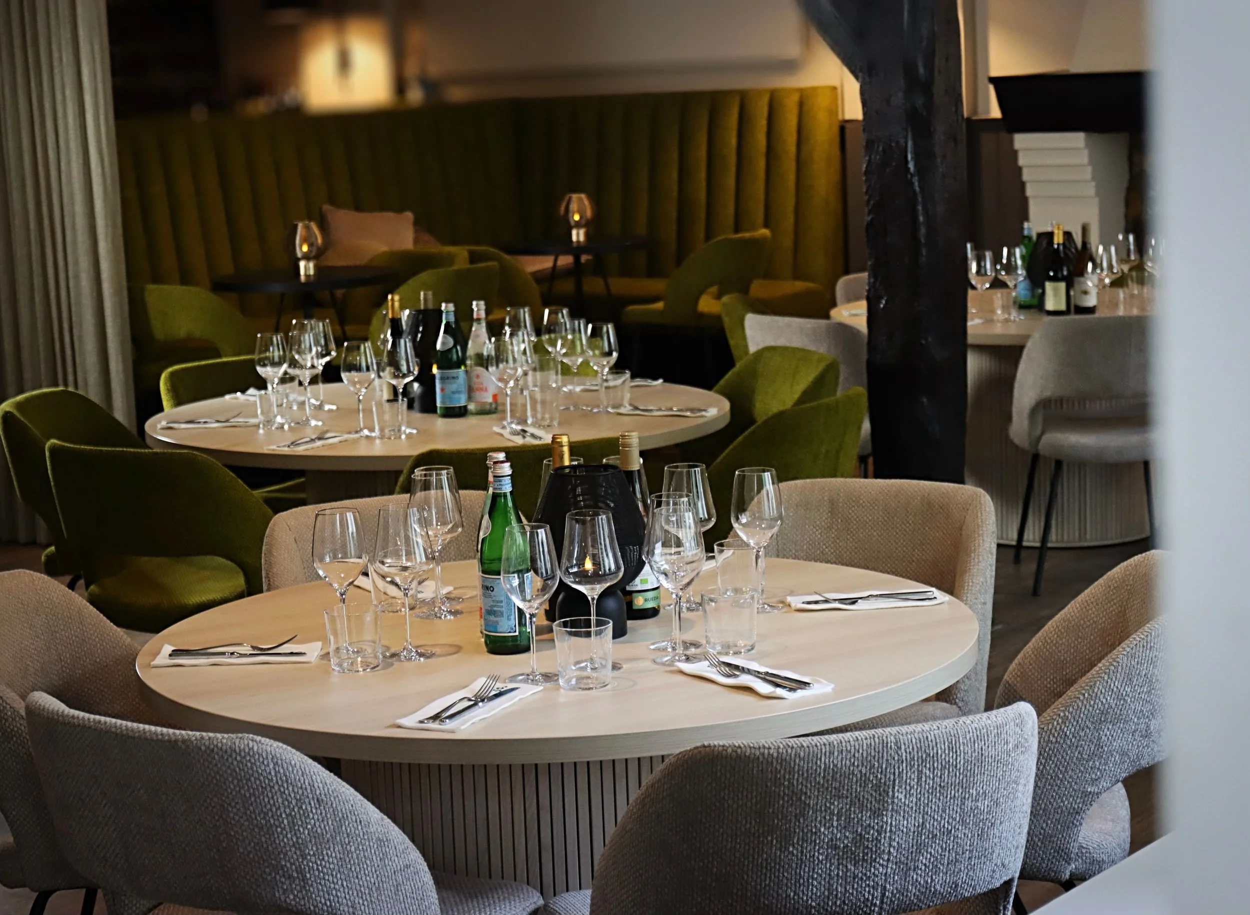 Een restaurant met ronde tafels gedekt met glaswerk, bestek en waterflessen, omgeven door comfortabele stoelen in neutrale en groene kleuren.