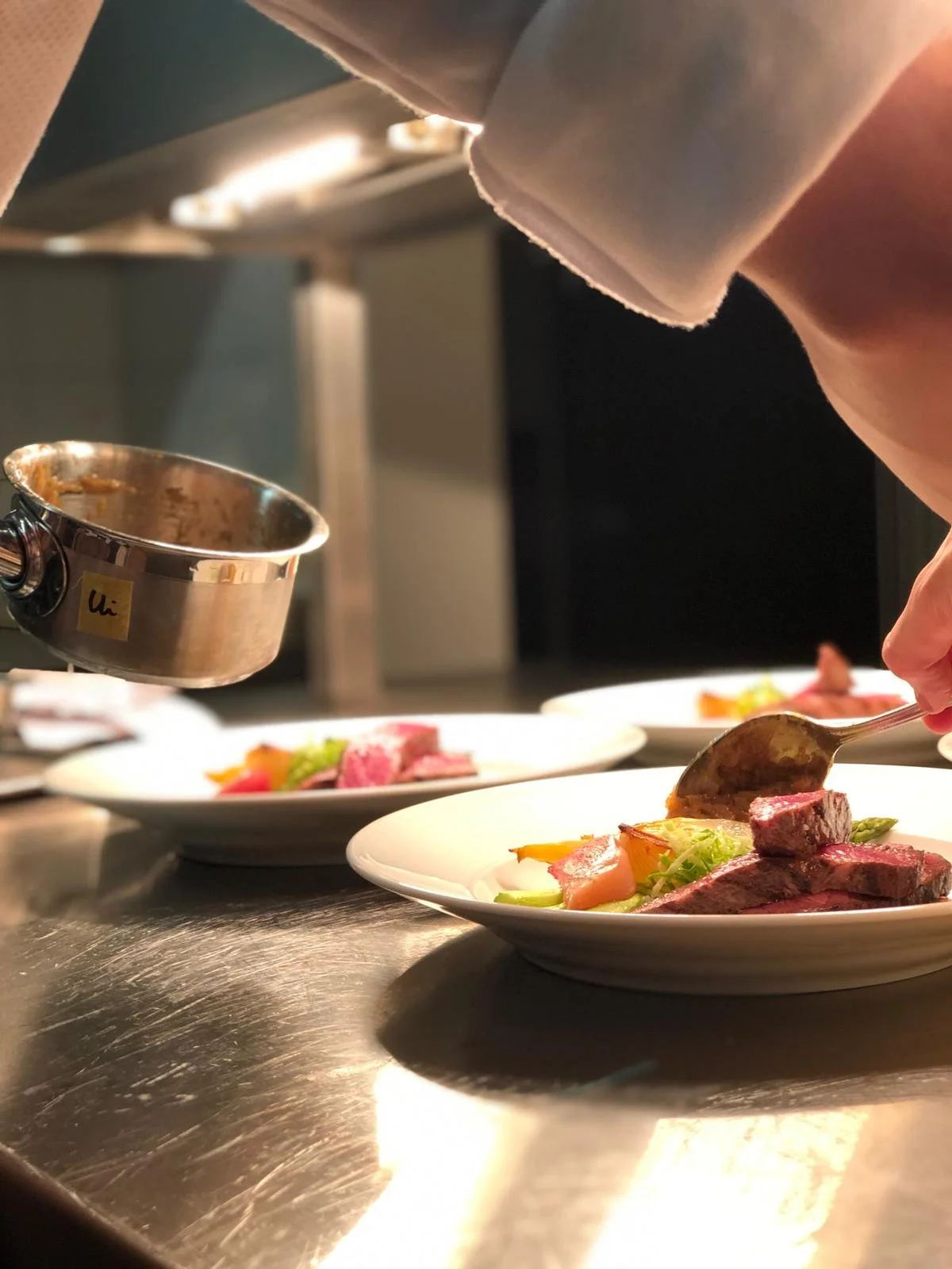 Chef serveert opbouwend gerecht met stukjes vlees en groenten op een wit bord in een professionele keuken.