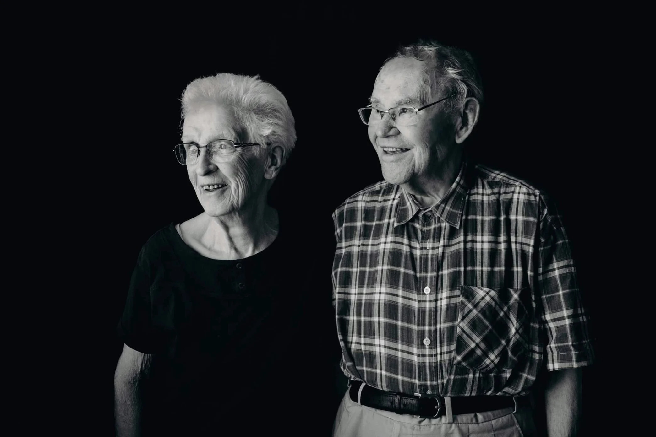 Un couple âgé souriant, avec un homme portant une chemise à carreaux et une femme avec des lunettes et un chemisier noir, sur fond sombre.