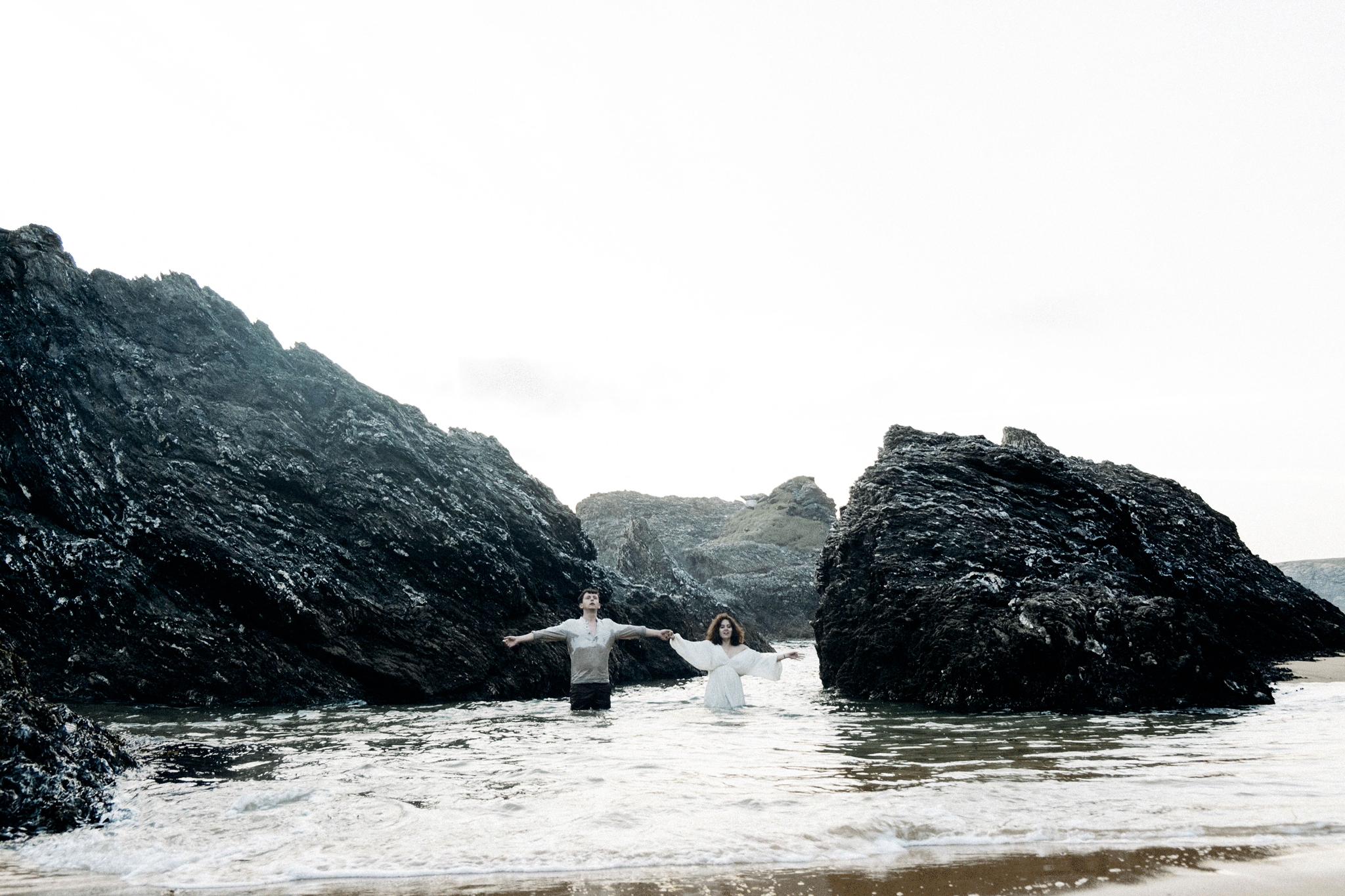 Deux personnes marchant dans l'eau entre des rochers à la plage, ciel blanc en arrière-plan.