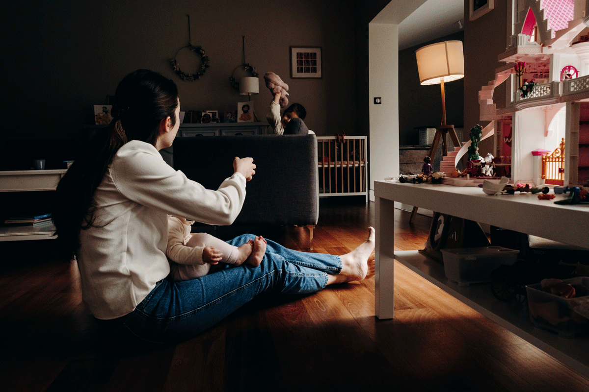 Une femme assise par terre tenant un bébé, avec une maison de poupées en plastique à droite et un enfant jouant avec une peluche en arrière-plan dans un salon.