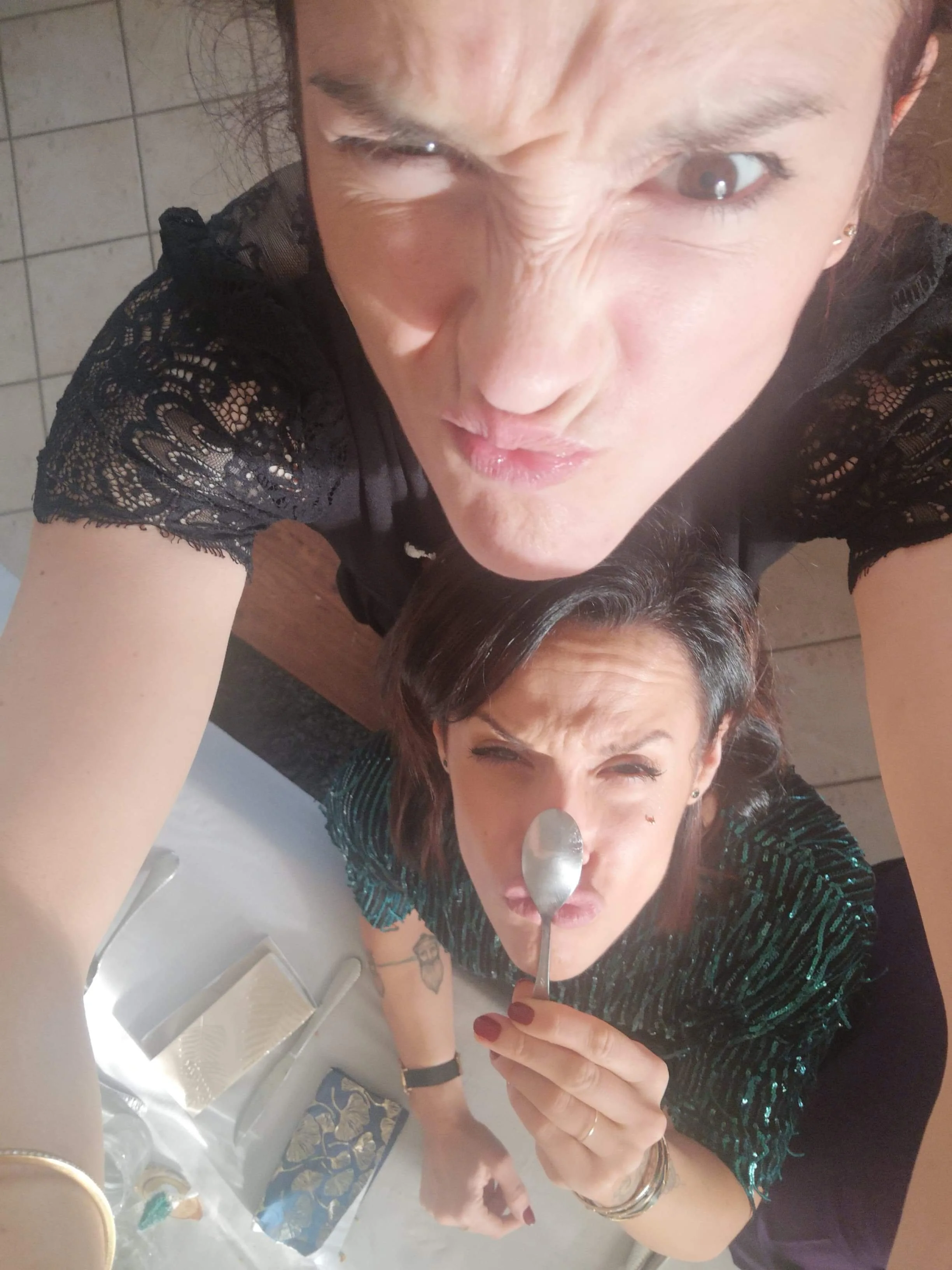 Deux personnes posant de manière amusante pour un selfie, l'une tenant une cuillère devant son visage. Elles sont dans une cuisine ou salle à manger éclairée par la lumière du jour, avec des ustensiles et serviettes visibles."
