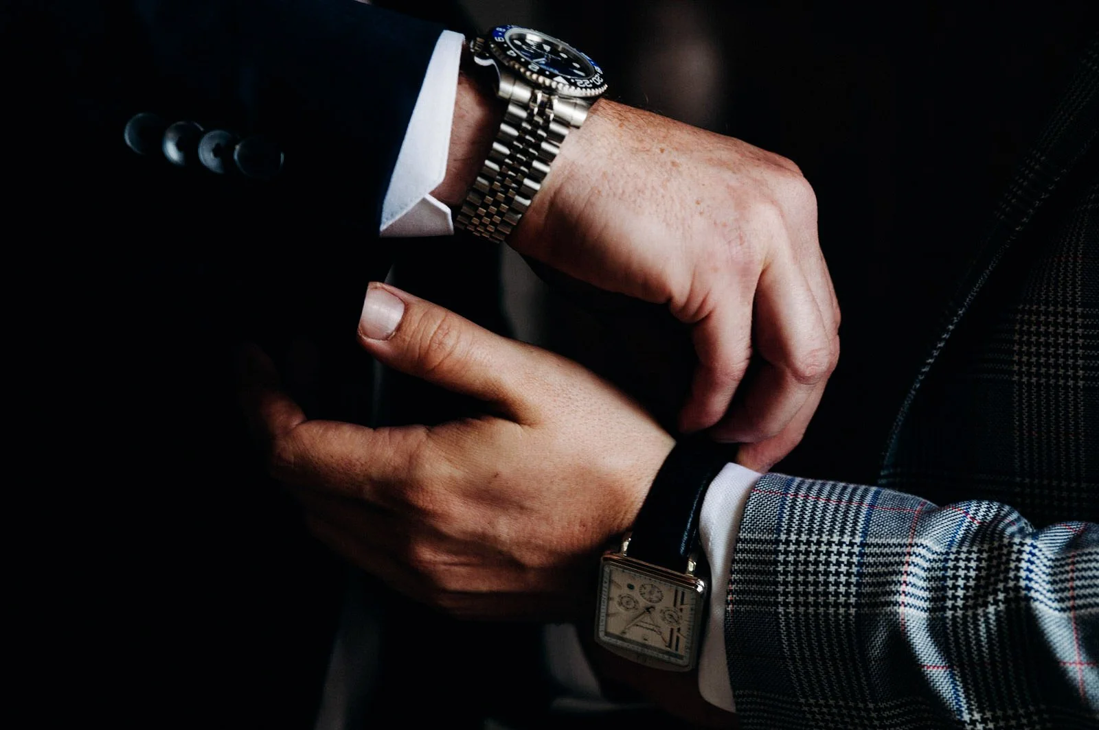 Deux hommes en costume ajustent leurs montres. L'un porte une montre en acier, l'autre une montre en cuir avec un cadran carré.