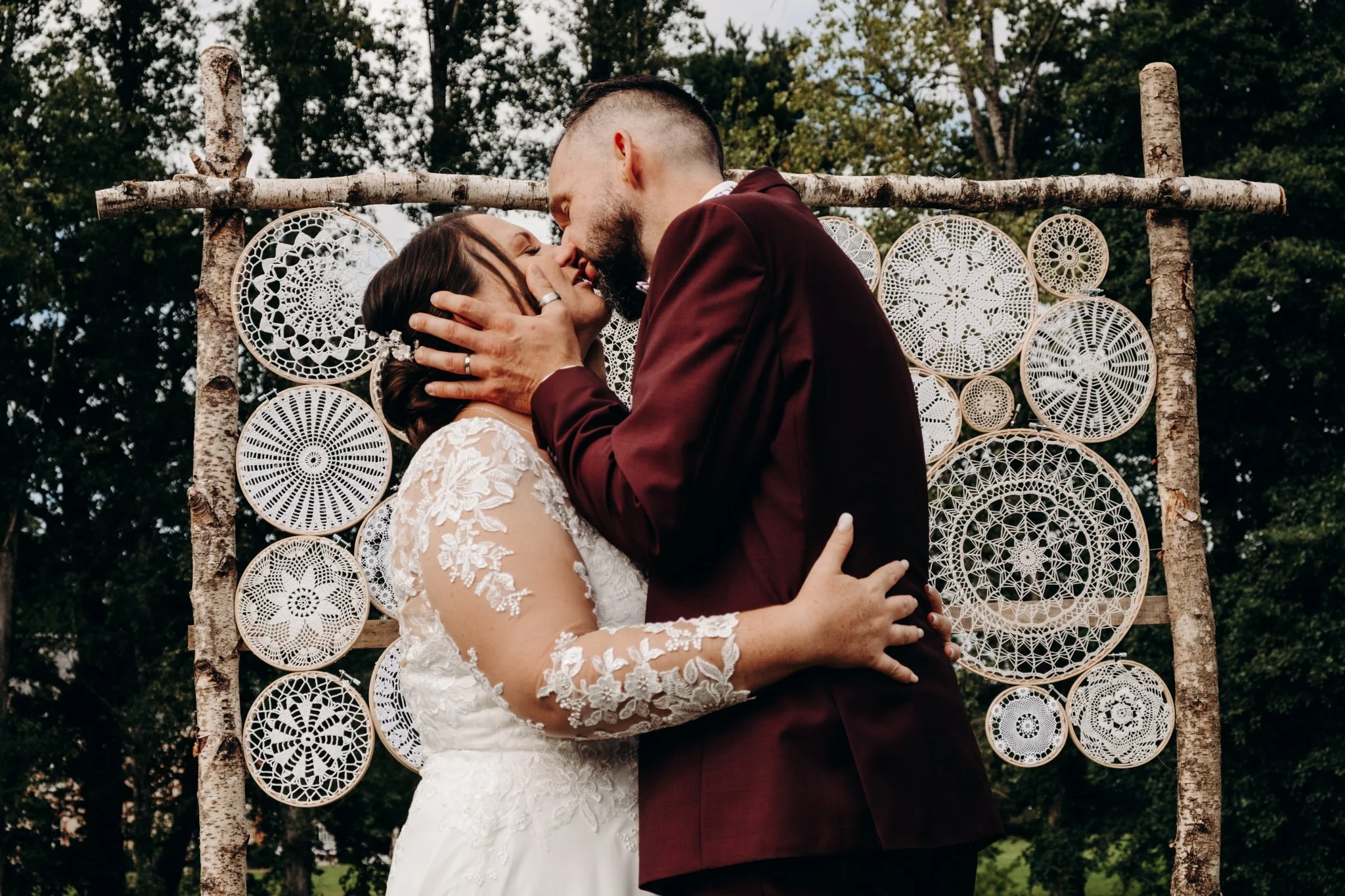 Un couple en tenue de mariage s'embrasse devant une décoration en dentelle et bois dans un cadre naturel.