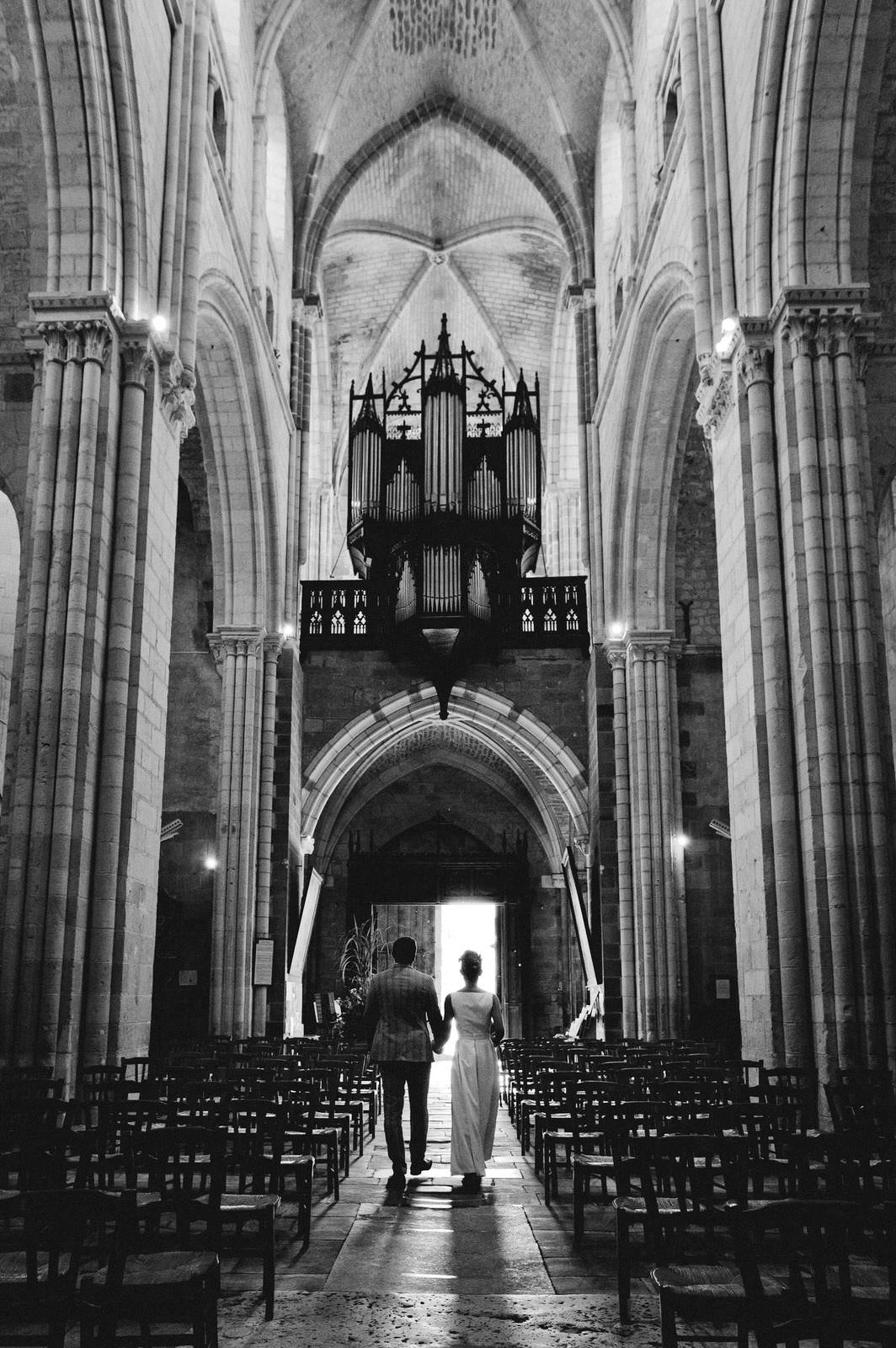 Un couple marche main dans la main dans une grande église, entouré de rangées de chaises vides et sous un plafond voûté.