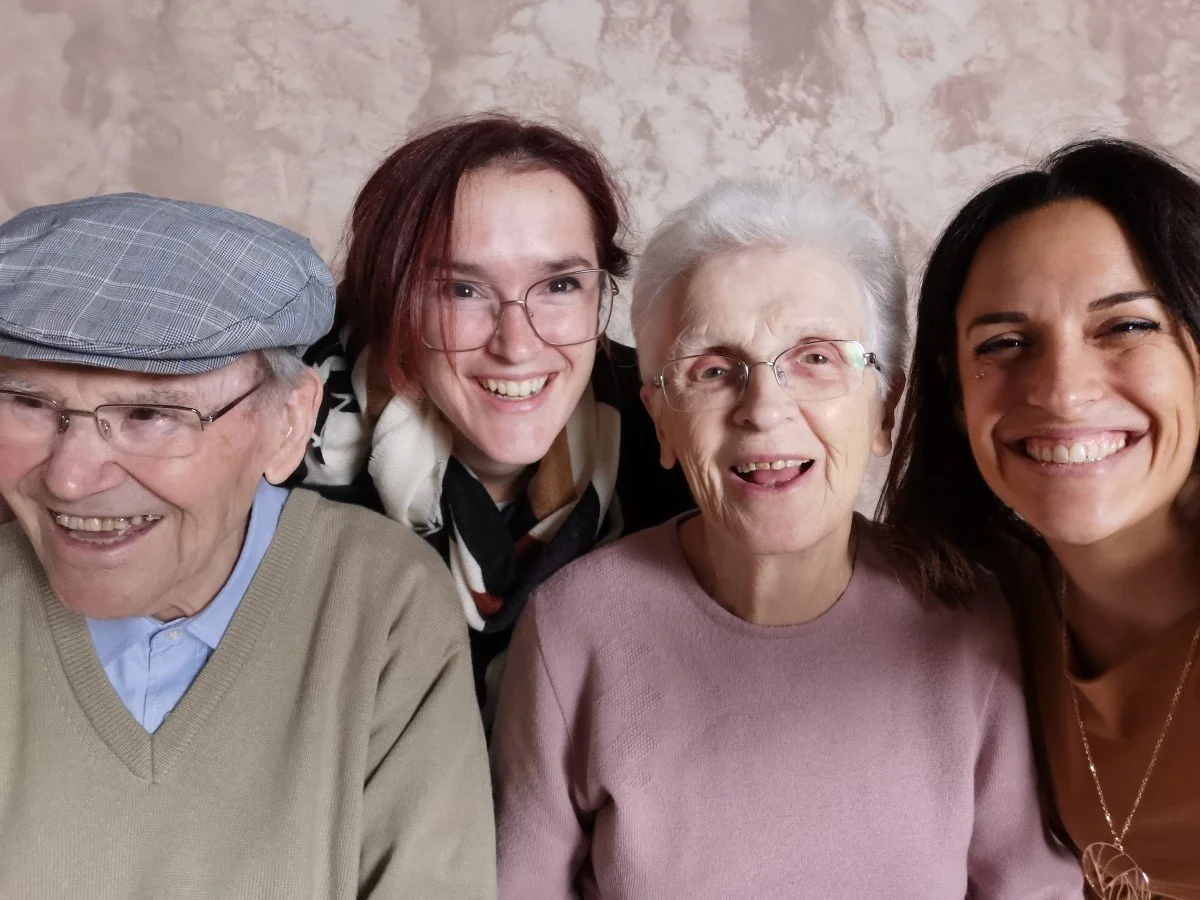Quatre personnes souriantes, deux hommes et deux femmes, posent ensemble pour une photo. L'homme à gauche porte une casquette et des lunettes. La femme à droite porte un collier.