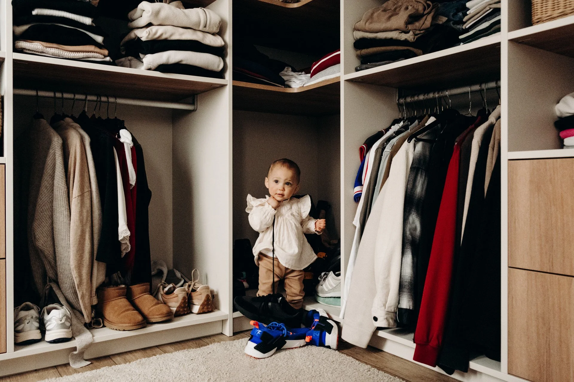 Un enfant se tient debout dans un placard rempli de vêtements et de chaussures, entouré de pulls pliés et de vestes suspendues.