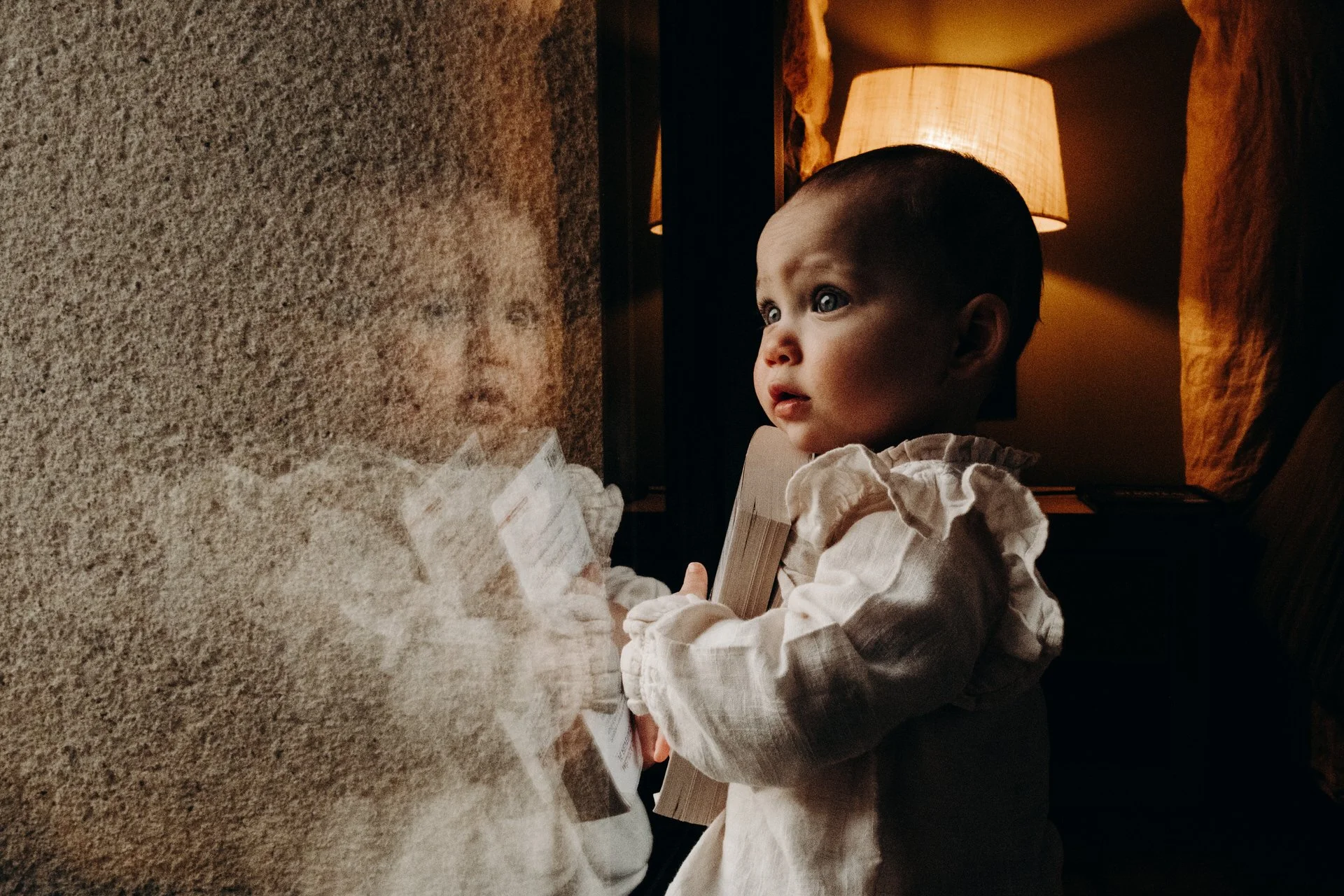 Bébé regardant par la fenêtre, reflet visible, luminaire en arrière-plan, ambiance chaleureuse.