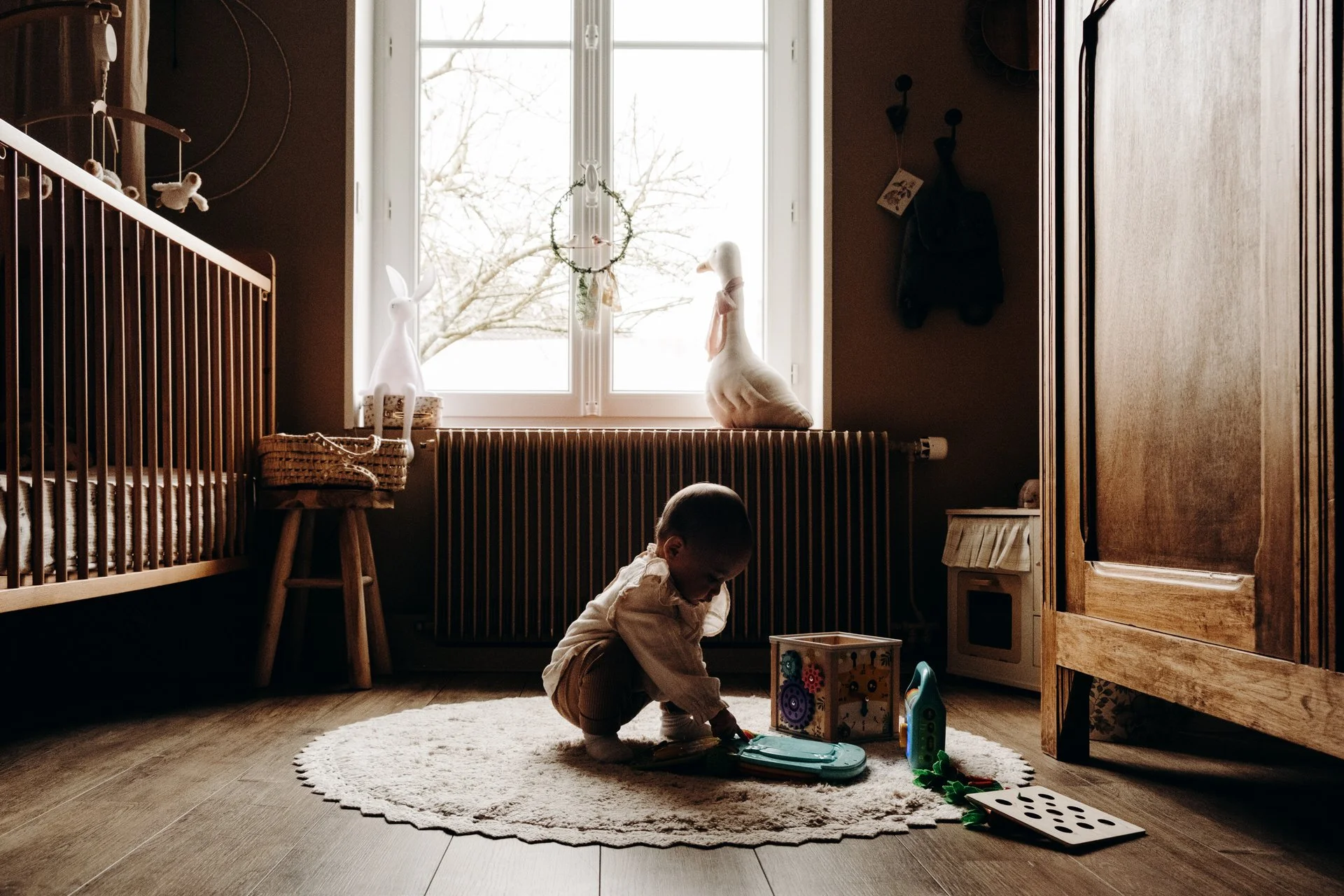 Bébé jouant dans une chambre avec jouets sur un tapis, éclairé par une fenêtre, lit et armoire en bois, décorations douces.