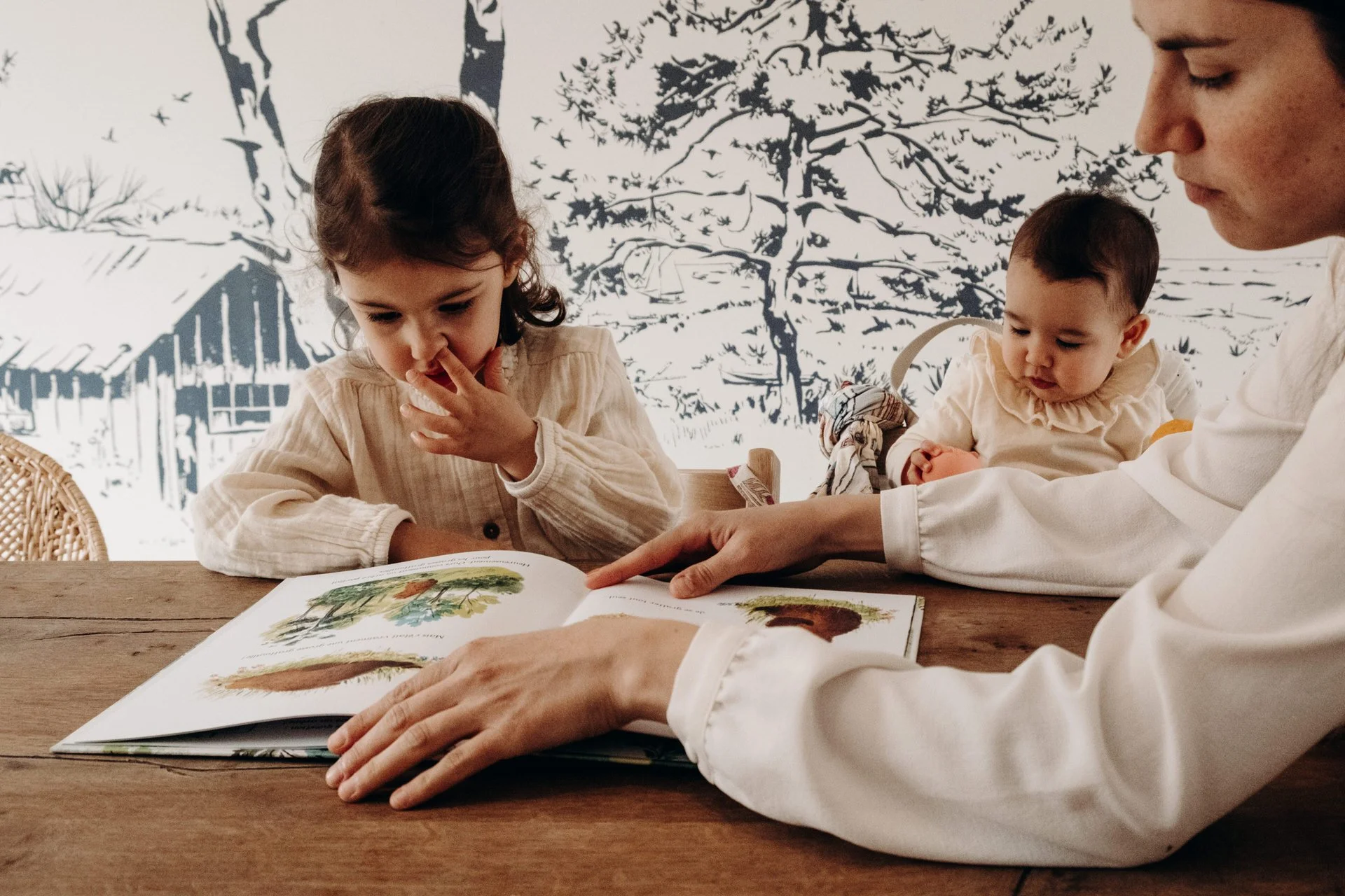Une femme et deux enfants lisant un livre illustré à une table en bois.
