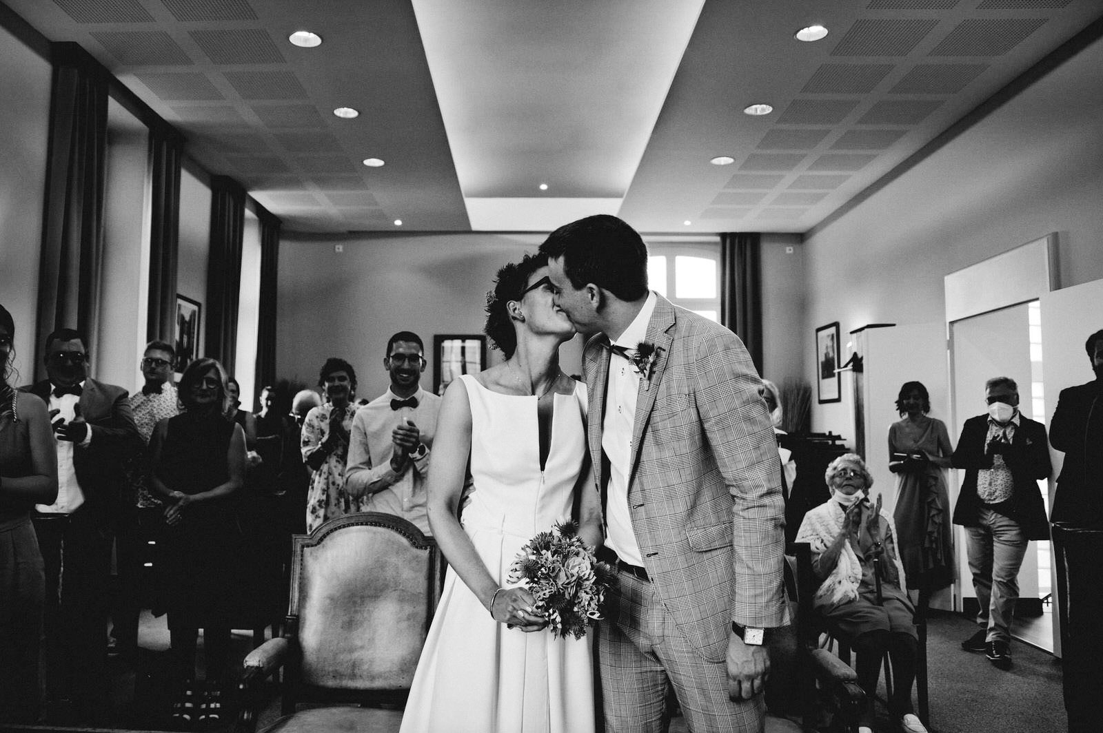 Un couple s'embrasse lors d'une cérémonie de mariage, entouré d'invités applaudissant, dans une salle élégante.