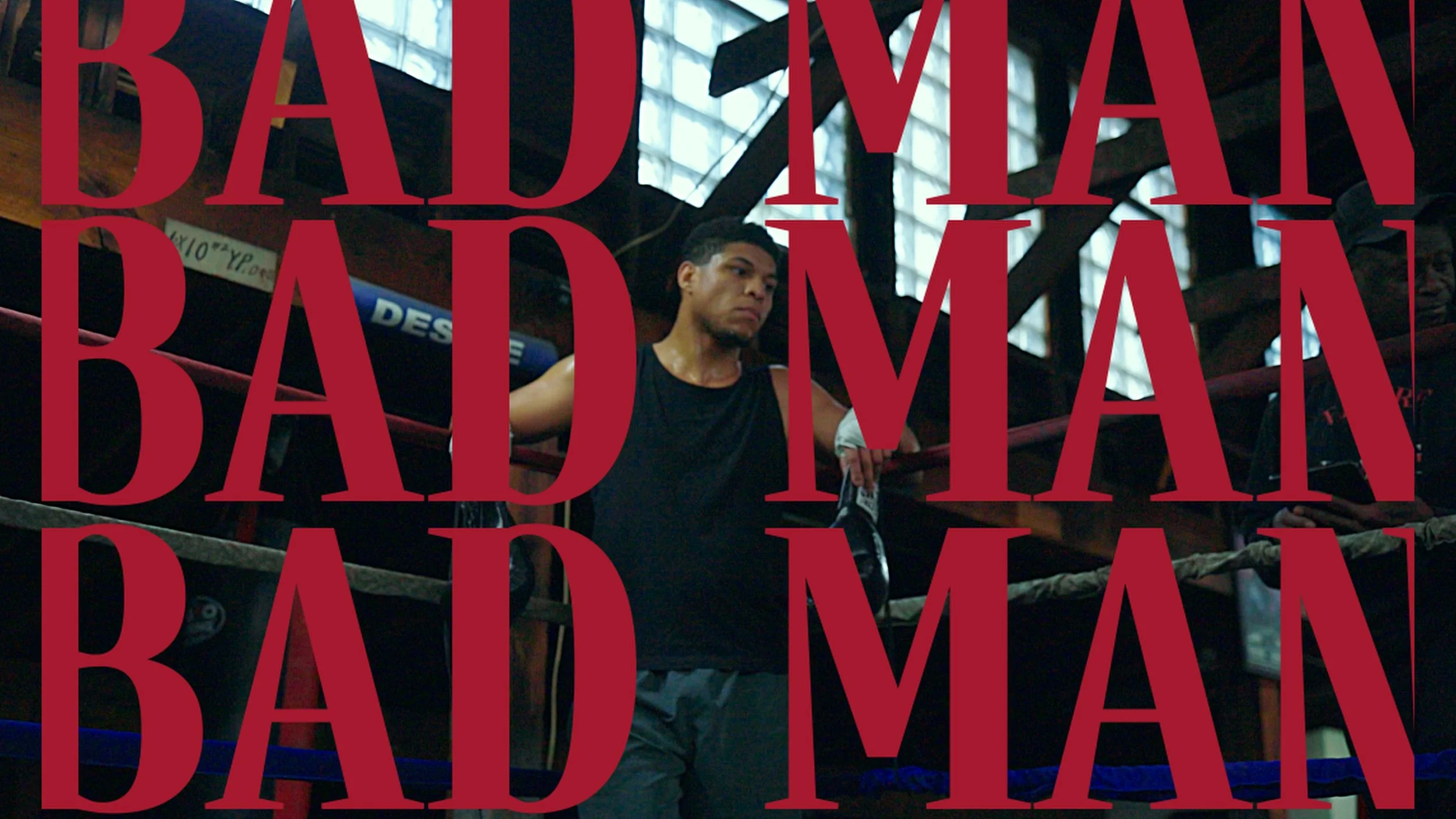 A young man wearing a black tank top and boxing gloves, standing inside a boxing ring with a serious expression, with large red text overlay reading 'BAD MAN' repeated multiple times.