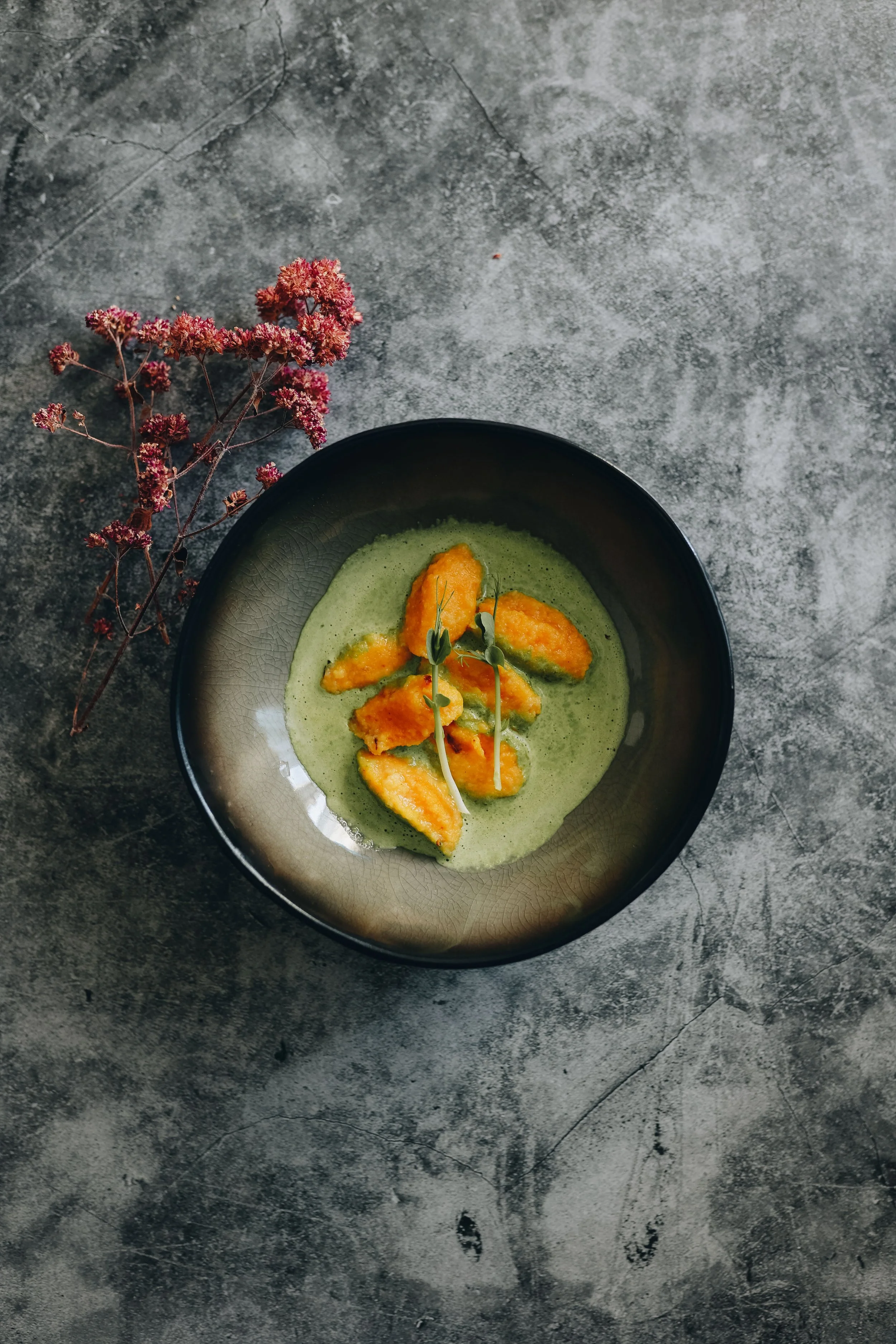 A black bowl containing green soup garnished with fried yellow pieces and microgreens, with dried pink flowers beside it on a textured gray surface.