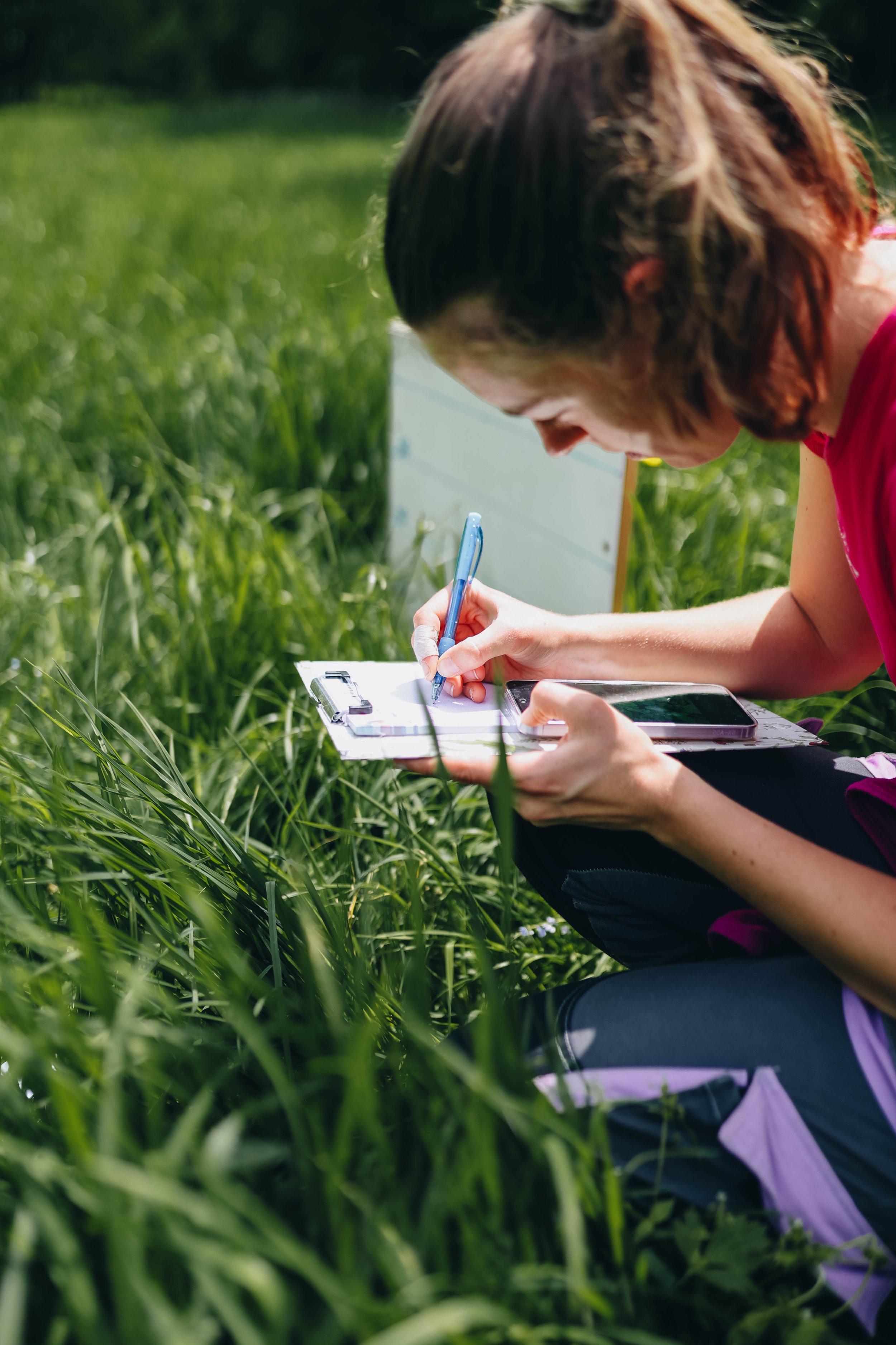 A woman with brown hair, wearing a red shirt, sitting in tall grass outdoors, writing on a clipboard with a pen, with a smartphone placed on her lap.