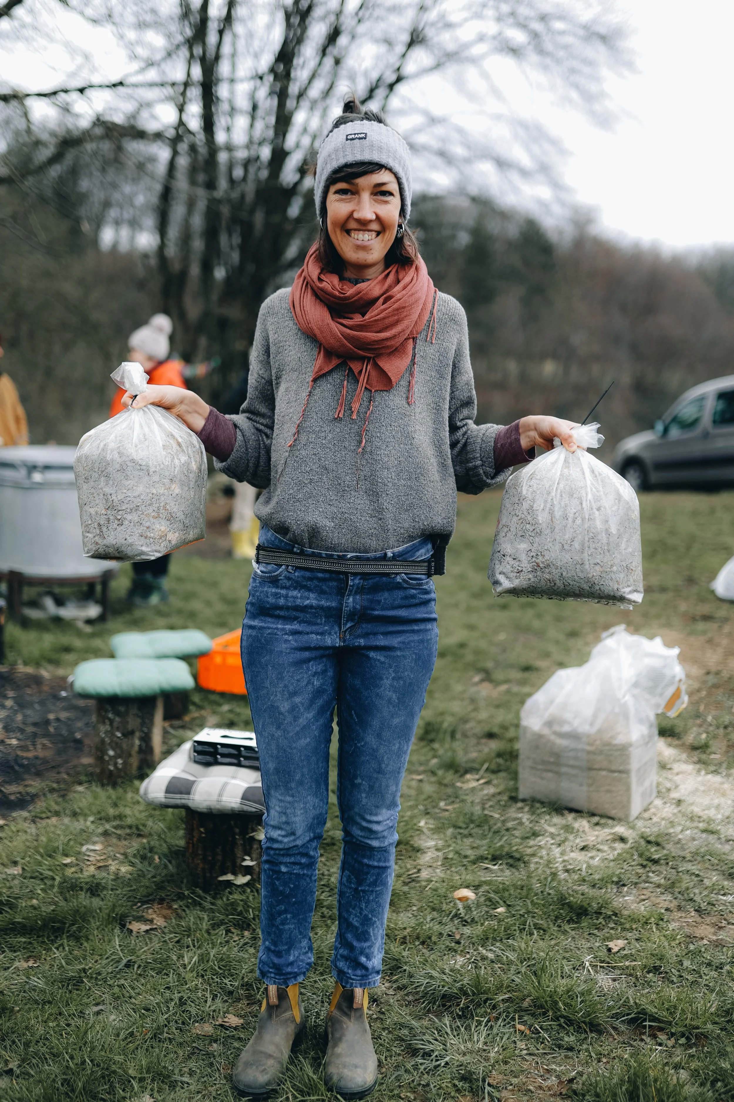 A woman outdoors smiling and holding two large plastic bags filled with compost or yard waste, with more bags and outdoor equipment visible in the background.