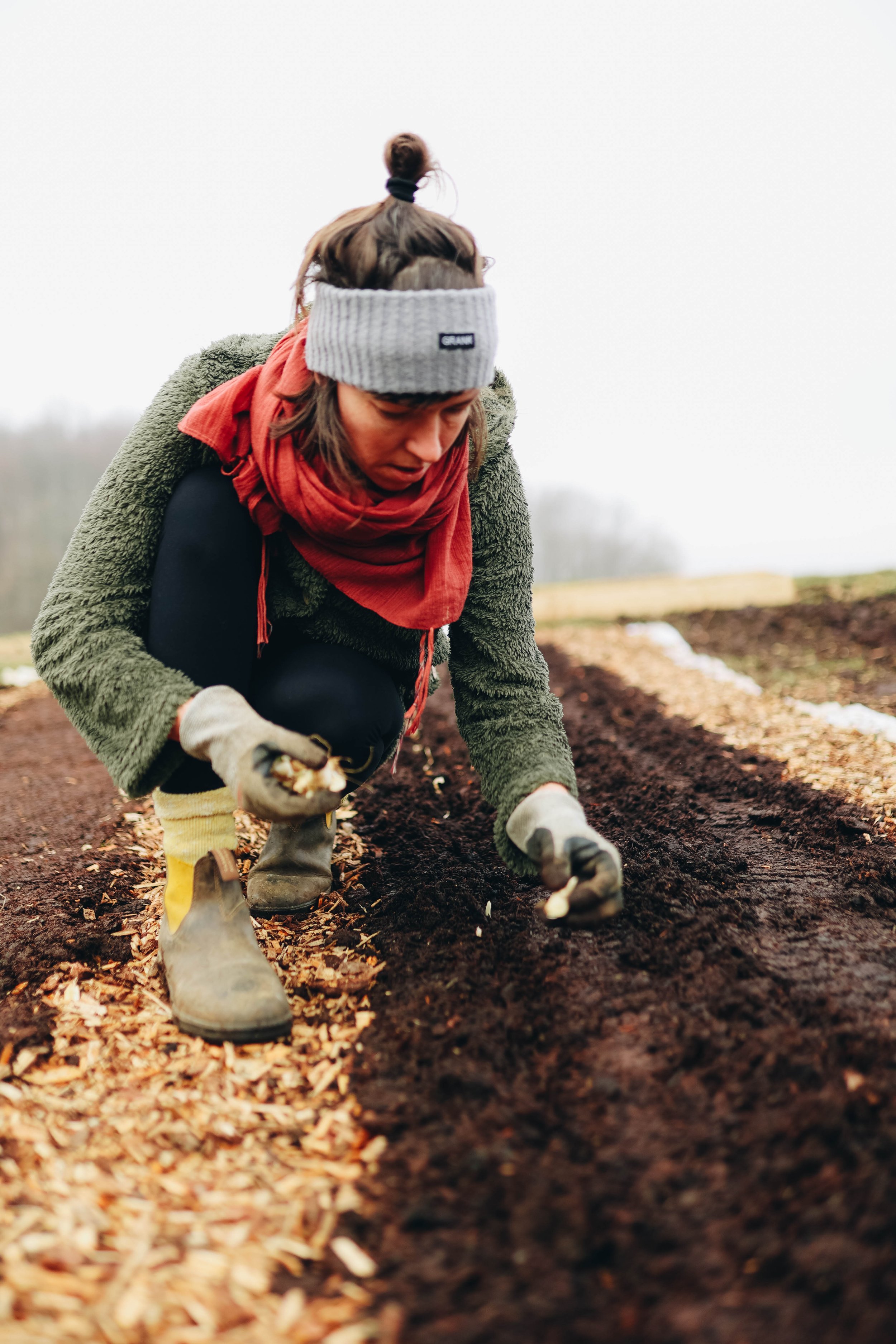 A person wearing a gray beanie, red scarf, green fuzzy jacket, black pants, and yellow boots, planting seeds in the soil of a garden bed on a cloudy day.