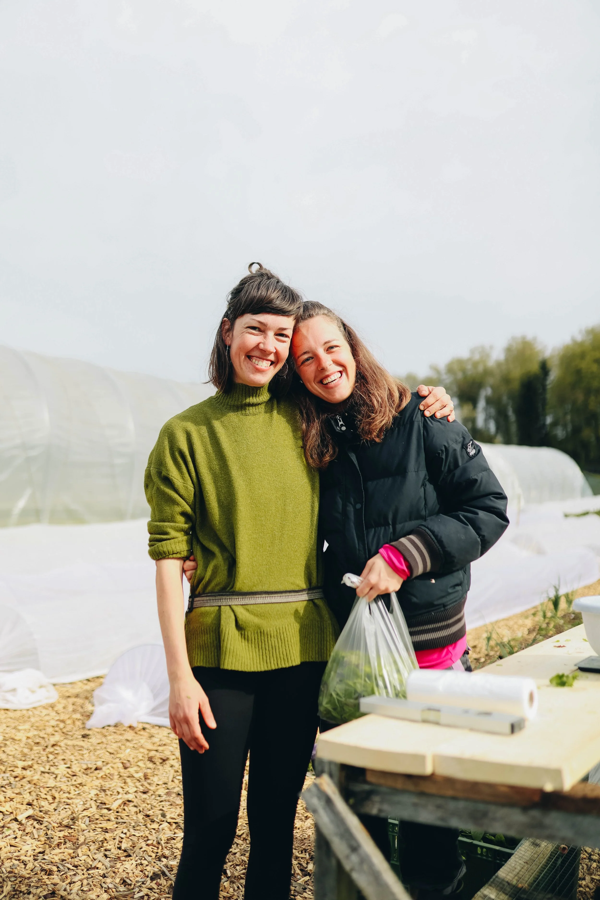 Two smiling women standing outdoors, one holding a plastic bag, in a garden area with greenhouses in the background.