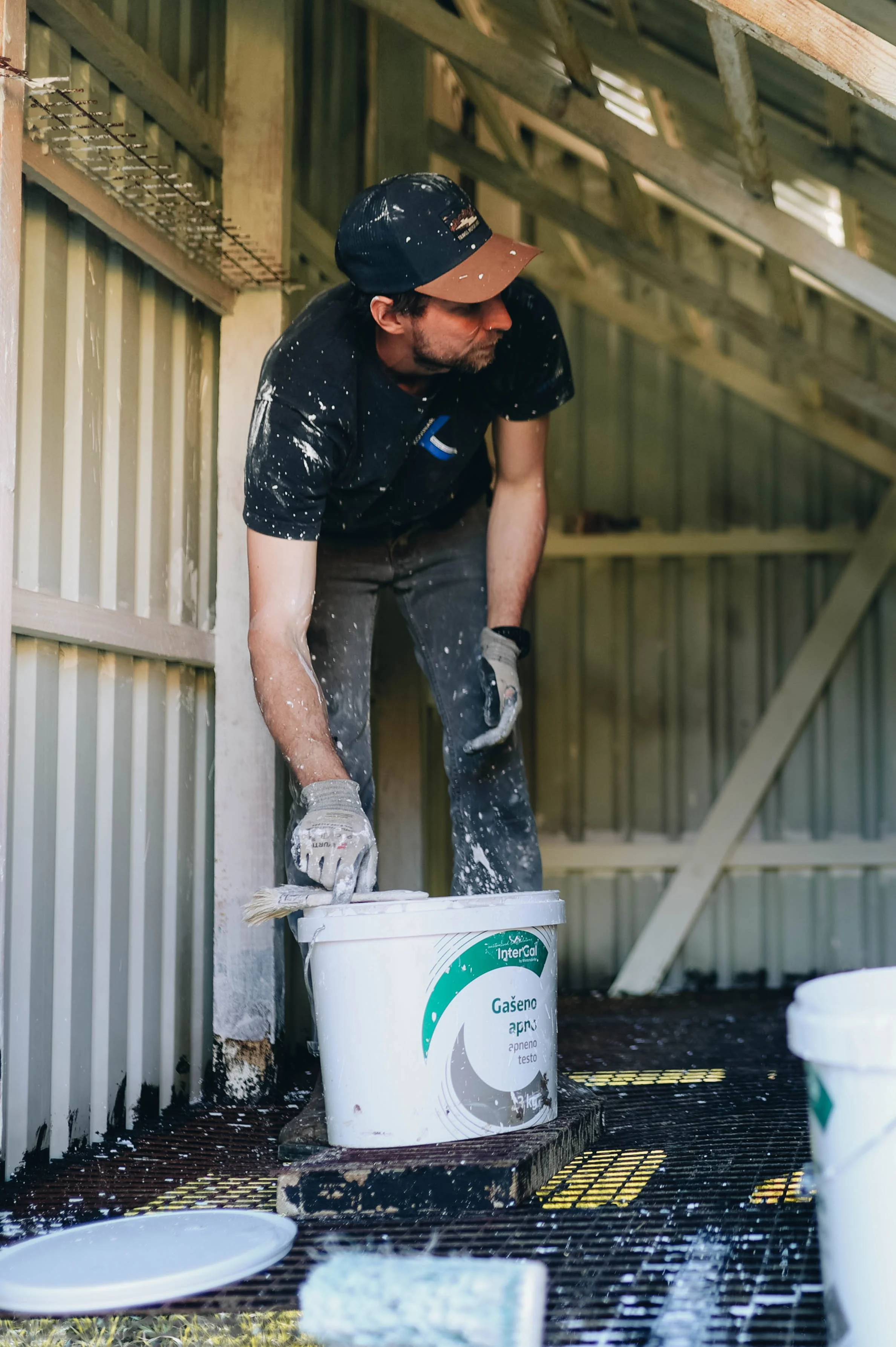 A man painting a wall inside a structure, using a roller brush, with paint splatters on his clothing and glove, standing on a black grated surface with buckets of paint nearby.
