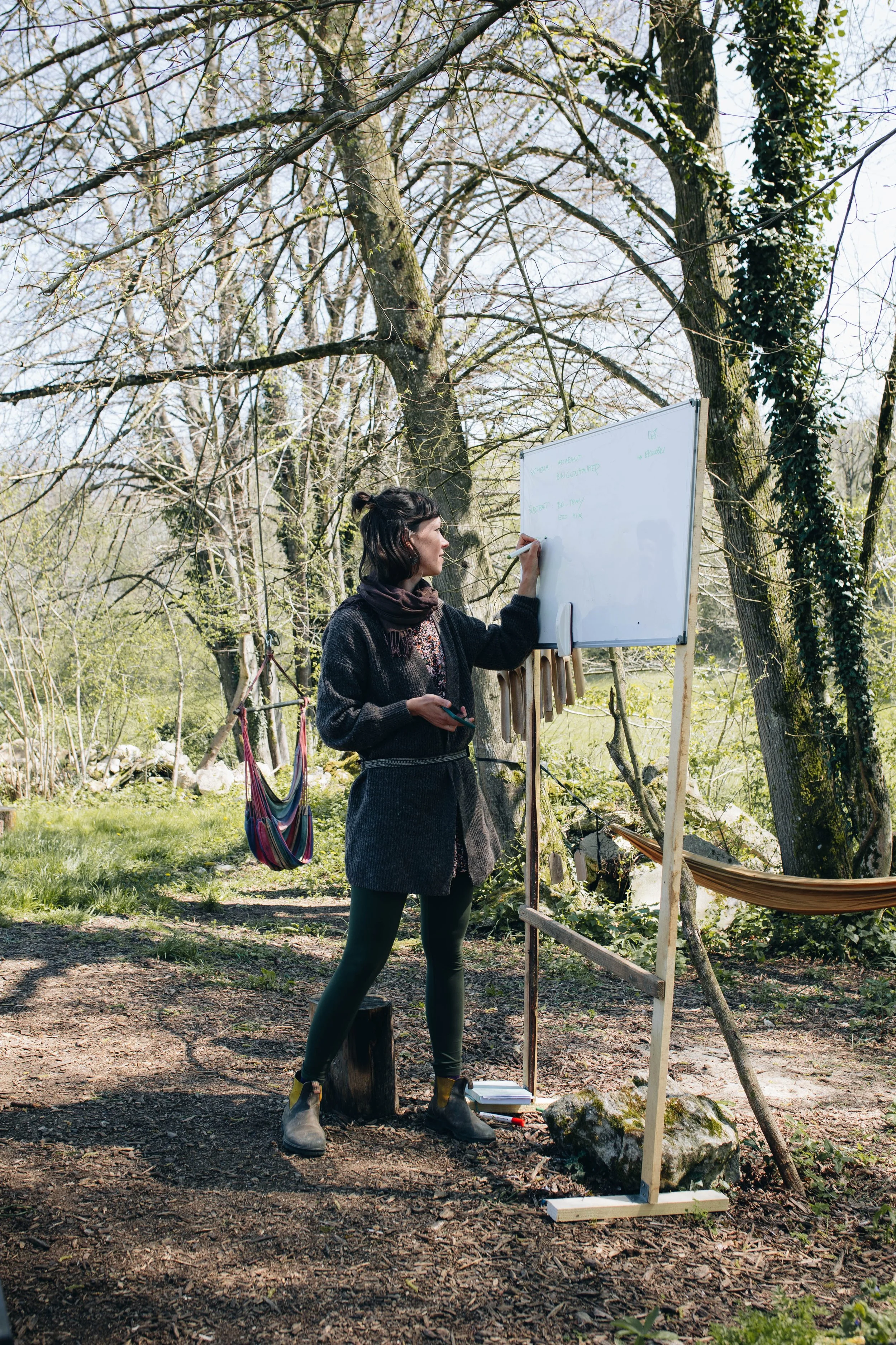 A woman writing on a whiteboard outdoors in a wooded area during daytime, with trees and a hammock in the background.