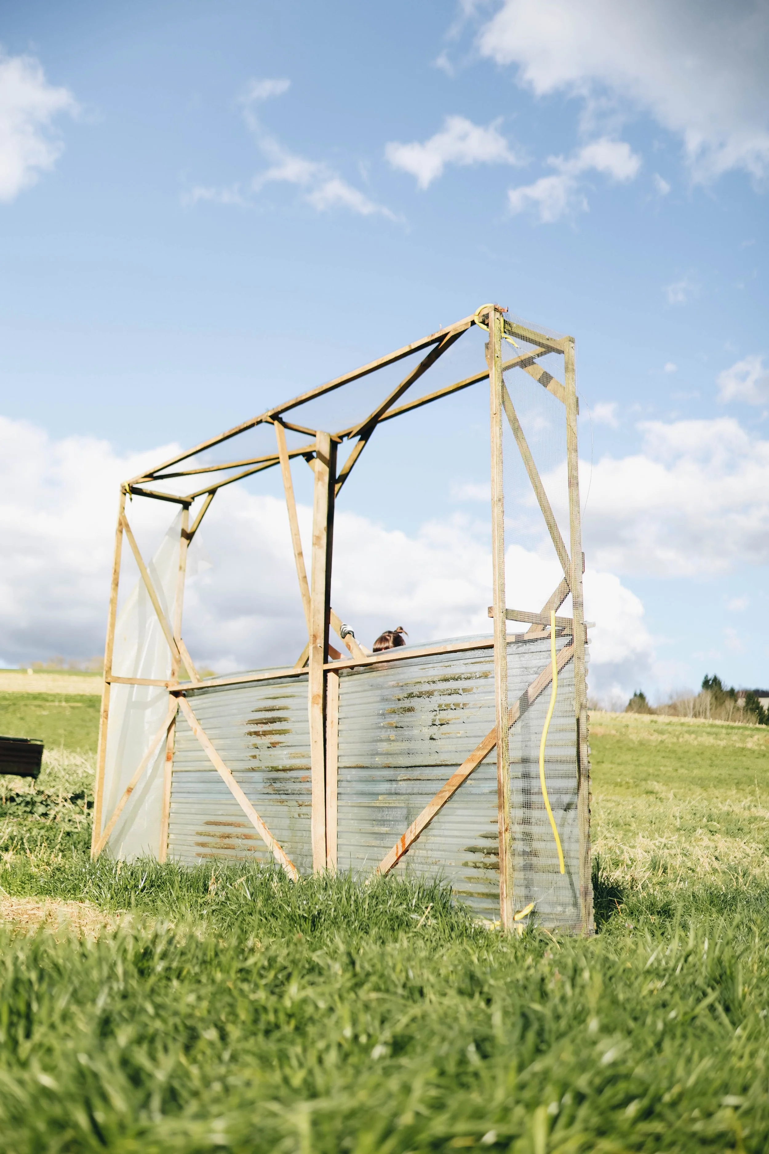 Unfinished wooden and metal structure in an open grassy field with a cloudy blue sky.